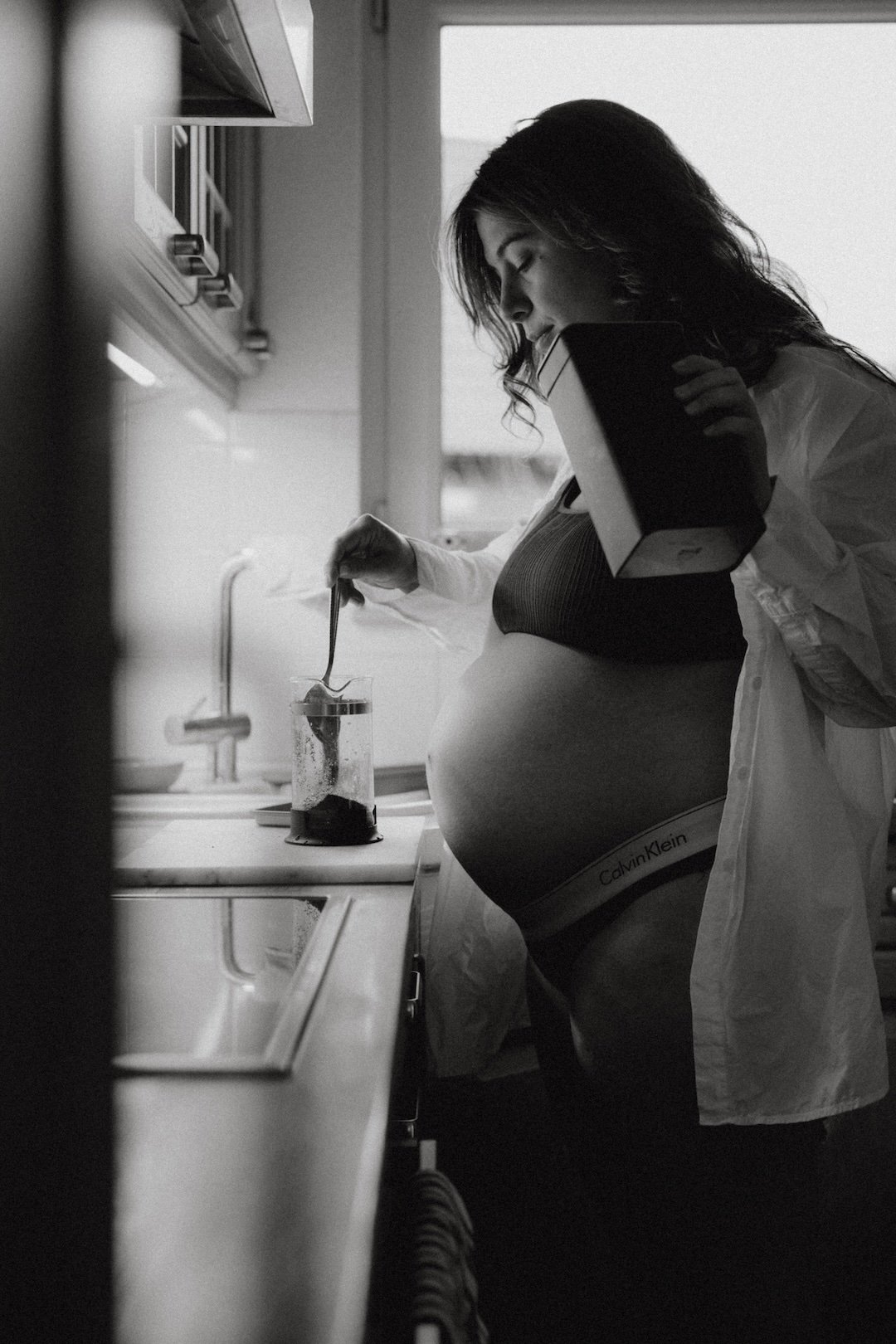 Schwangere Frau in der Küche, die Zucker in eine Küchenmaschine gibt, während sie ein Buch hält, und im Vordergrund ein Tuch hängt.