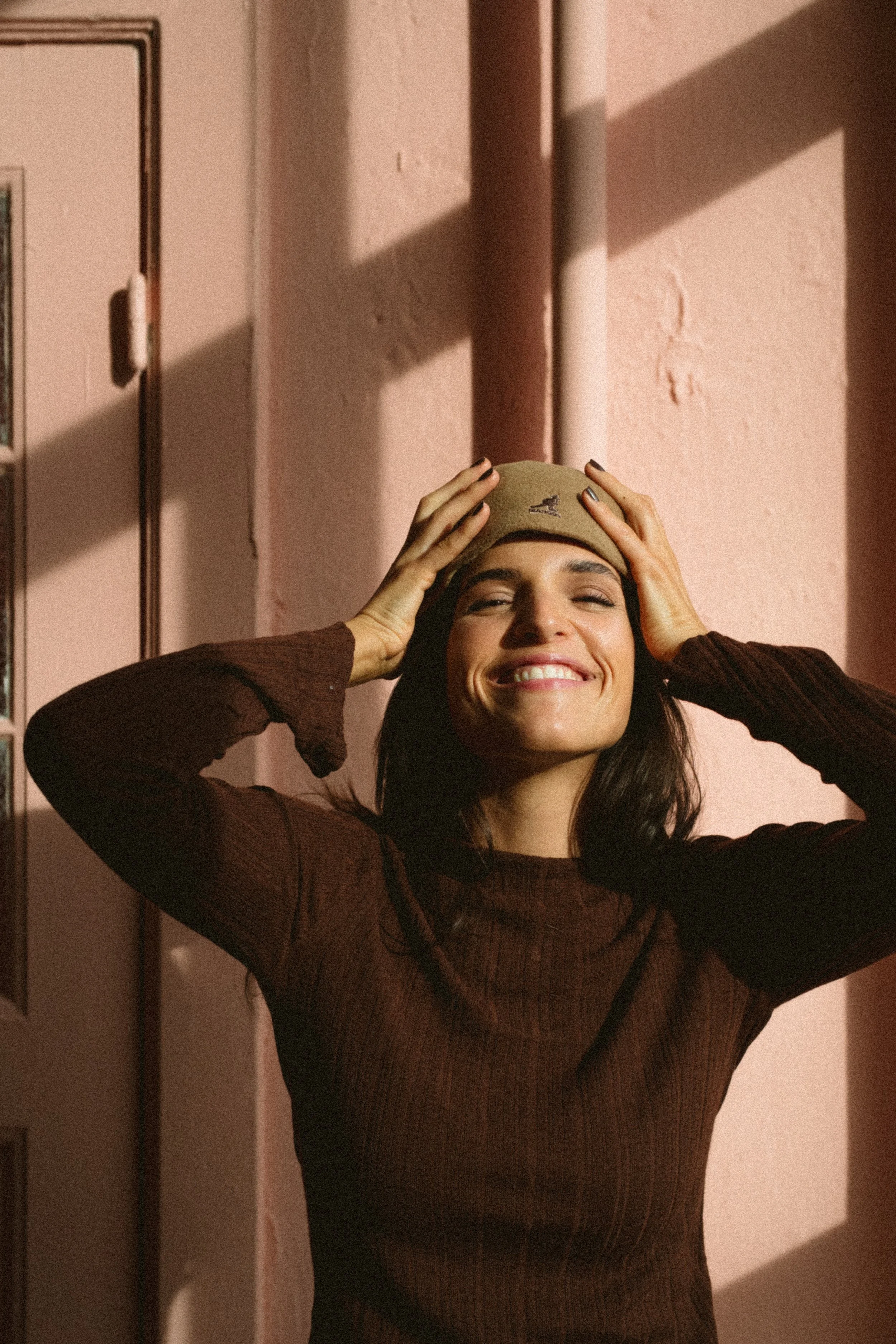 Lächelnde junge Frau trägt eine beige Mütze und einen braunen Pullover, steht vor einer pinkfarbenen Wand mit Schatten