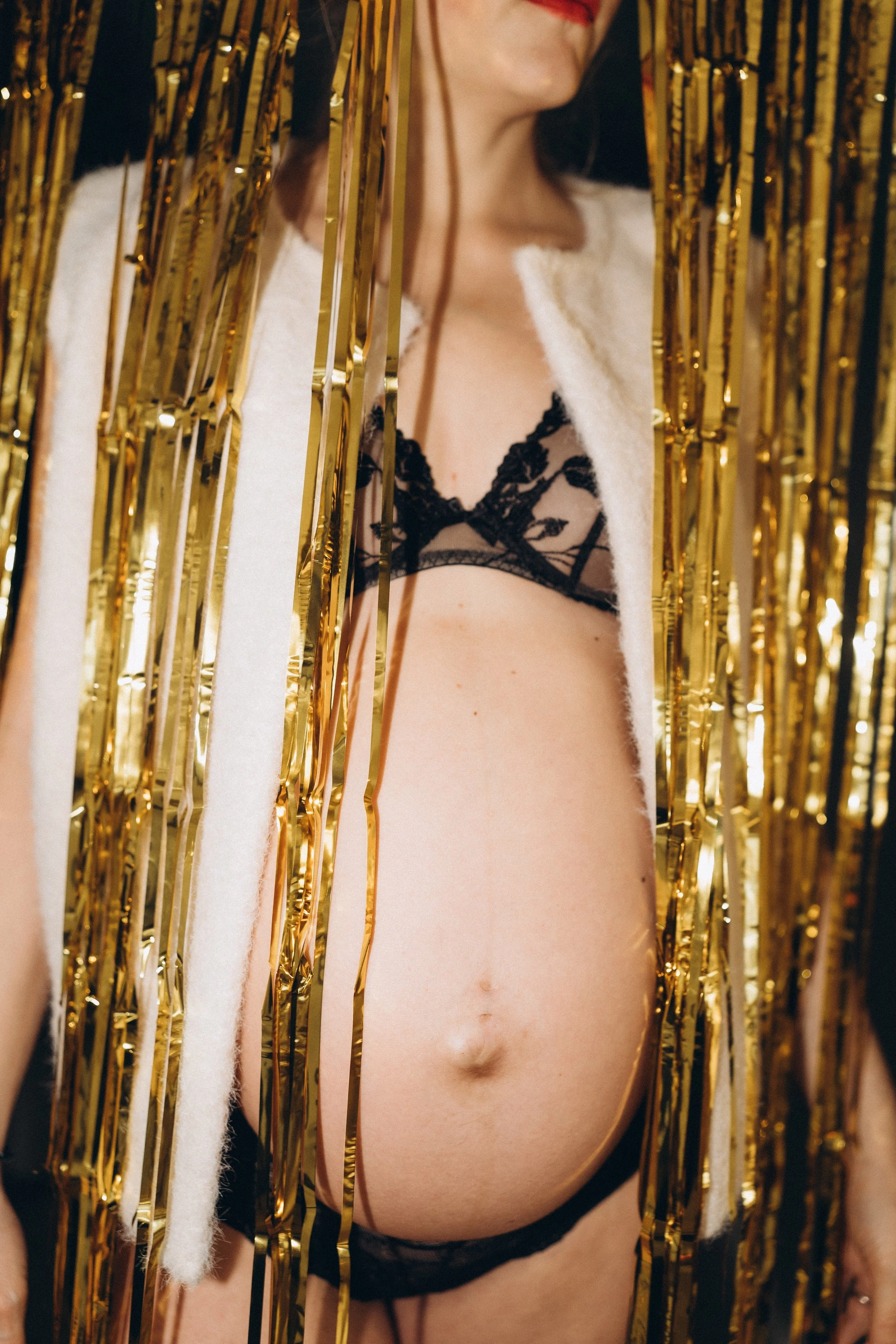 Eine schwangere Frau in schwarzer Spitze-Unterwäsche, teilweise verdeckt von goldener Dekoration und Weißstoff, auf einem festlichen Hintergrund.