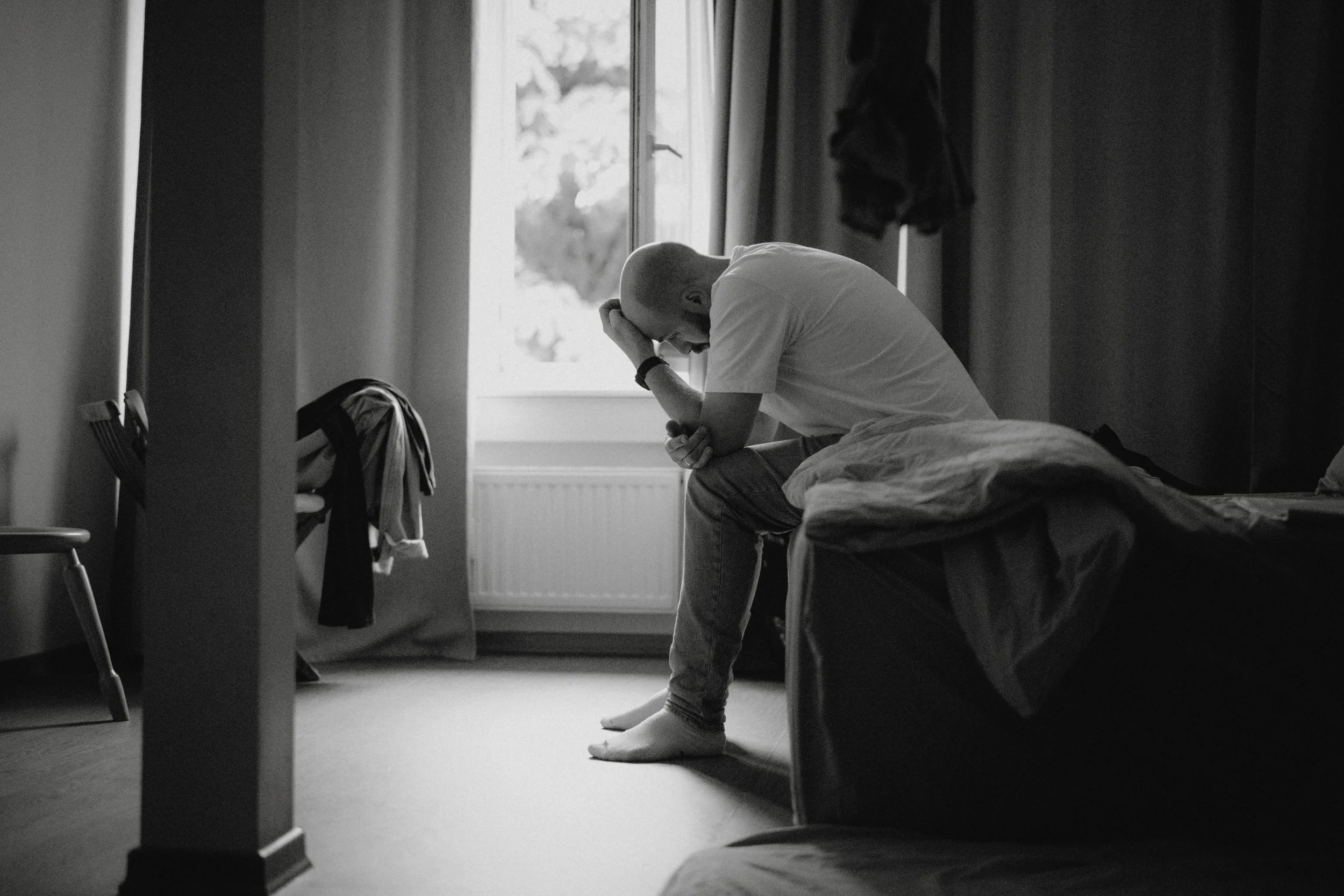 Mann sitzt auf einem Bett, den Kopf in der Hand, in einem hell erleuchteten Raum, schwarz-weiß