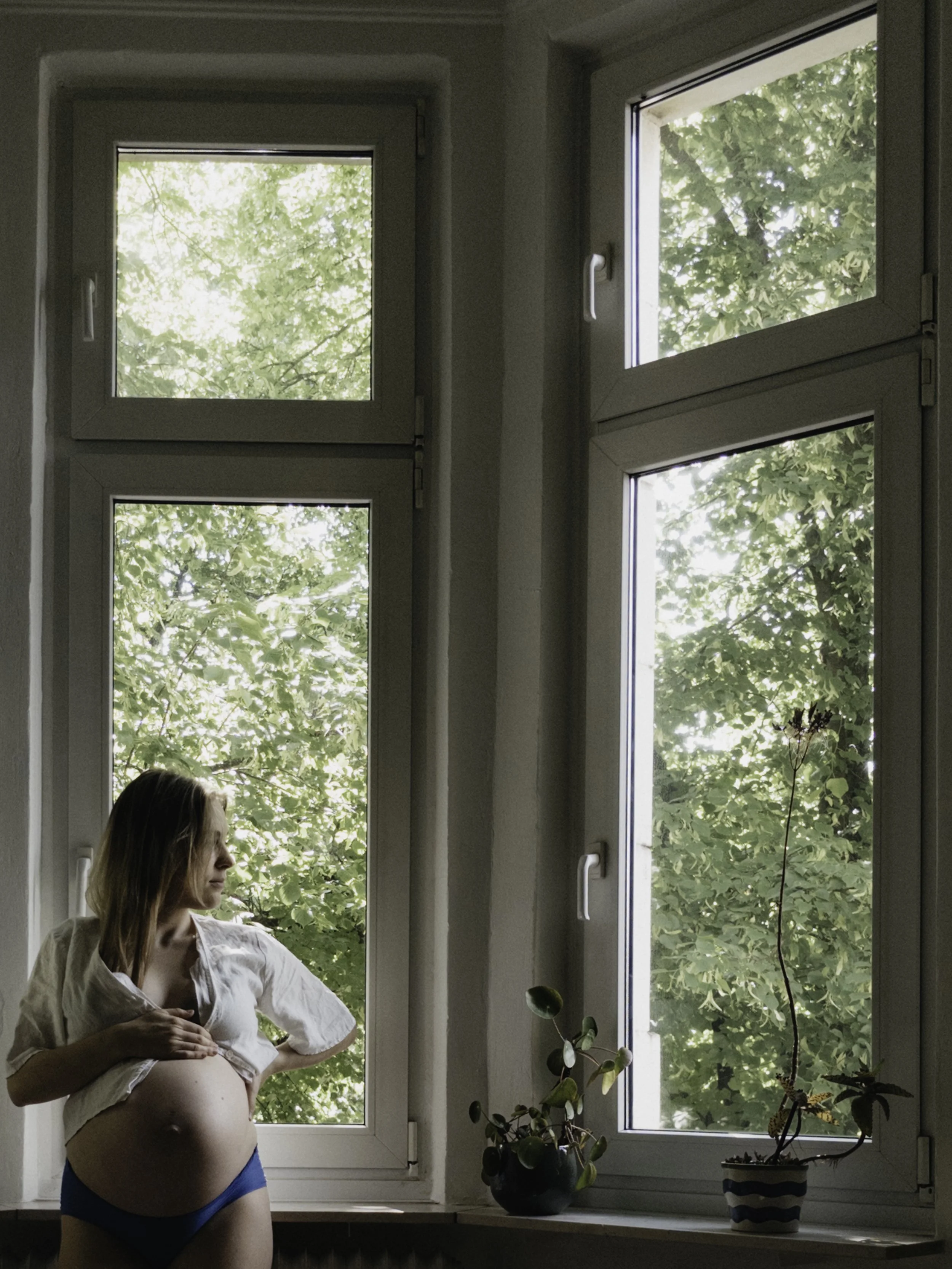 Eine schwangere Frau steht vor einem Fenster, das Blick auf grüne Bäume bietet, in einem Raum mit Pflanzen auf der Fensterbank.