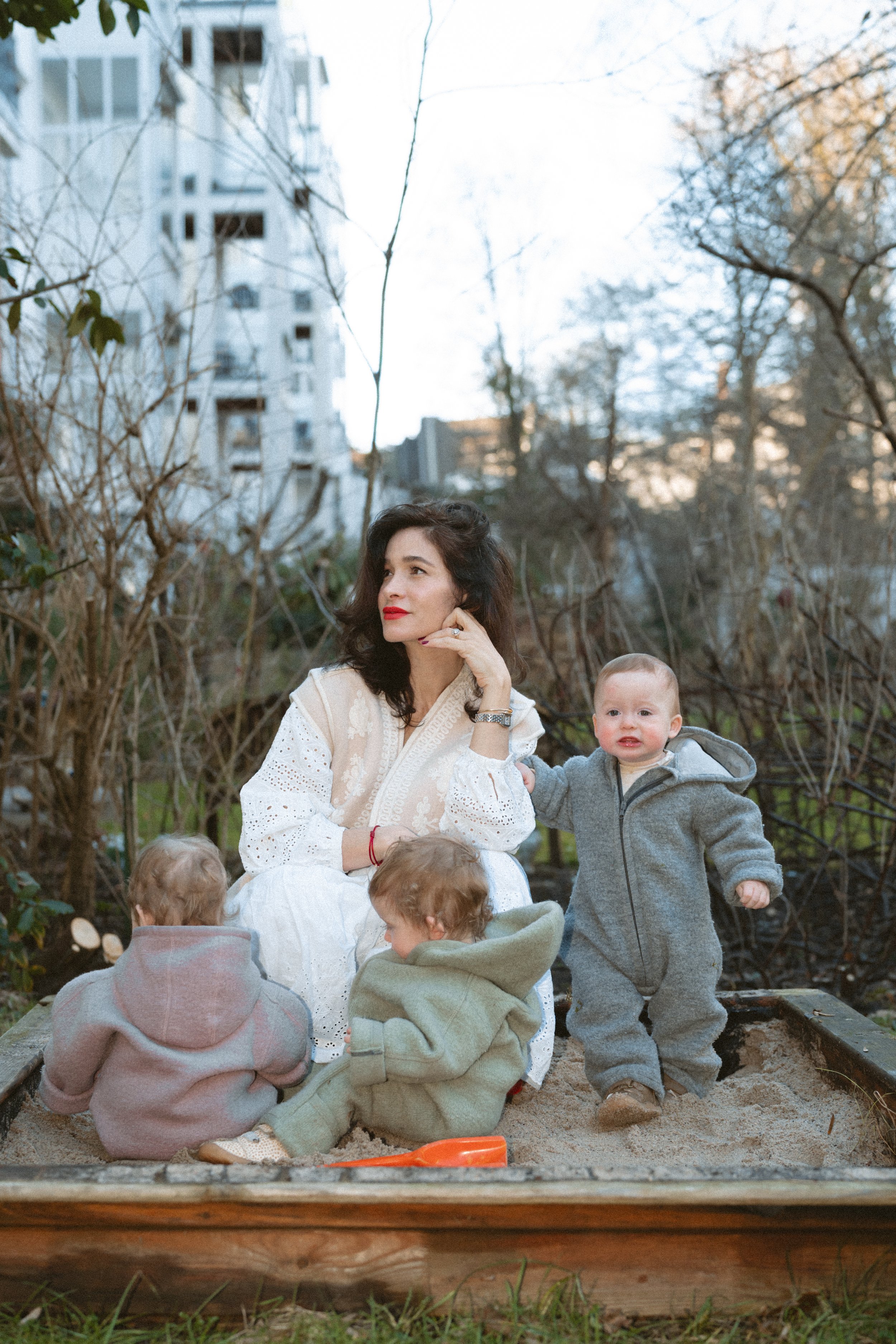 Frau mit drei kleinen Kindern spielt im Sandkasten in einem Garten, umgeben von Büschen und Bäumen, im Hintergrund sind Gebäude sichtbar.