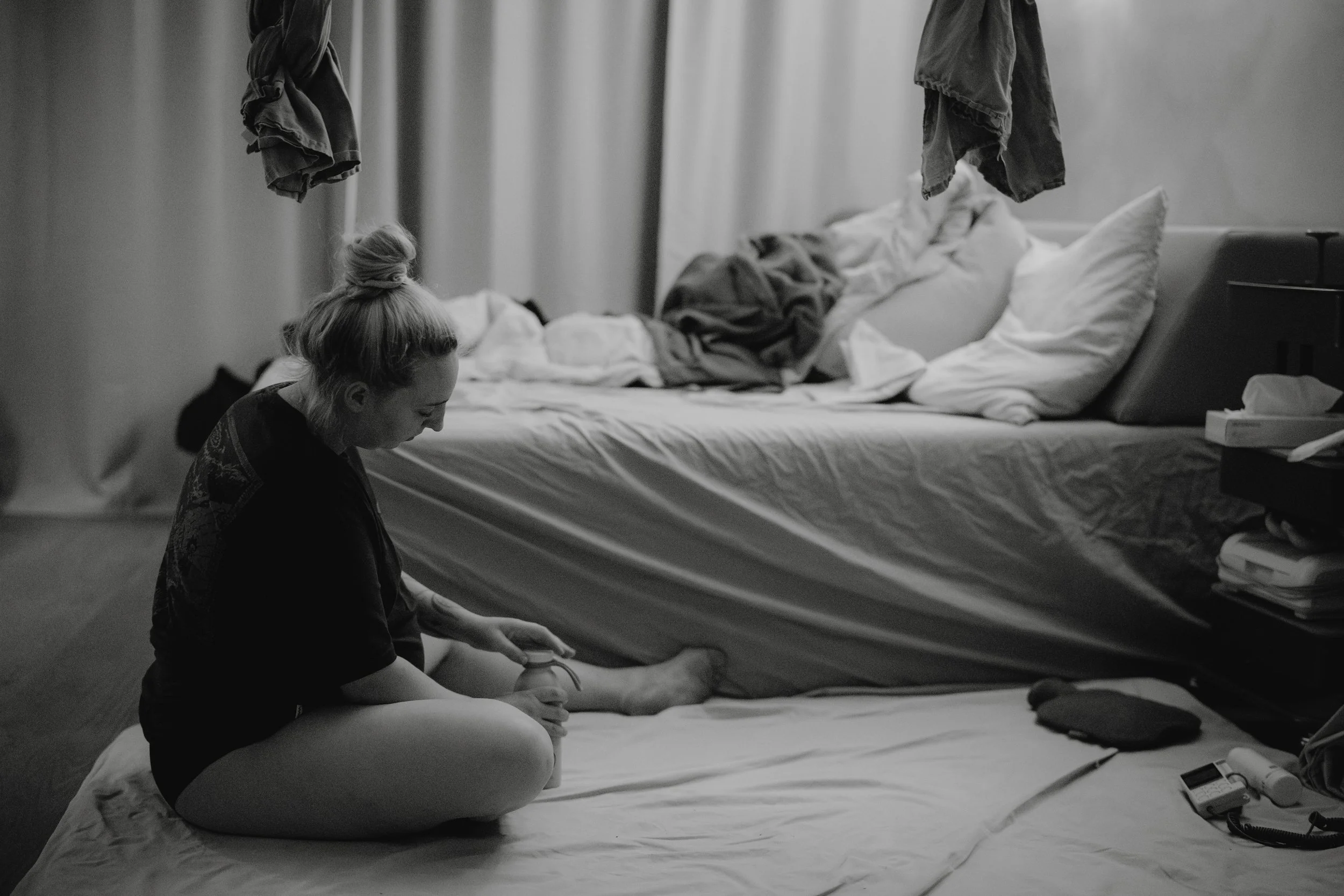 Eine Frau sitzt auf dem Boden eines unordentlichen Schlafzimmers, während sie mit einem elektrischen Rasierer an ihrem Bein rasiert, neben ihr liegt eine Matte oder Decke, und das Bett dahinter ist ungeordnet mit herumliegenden Kissen und Bettwäsche.