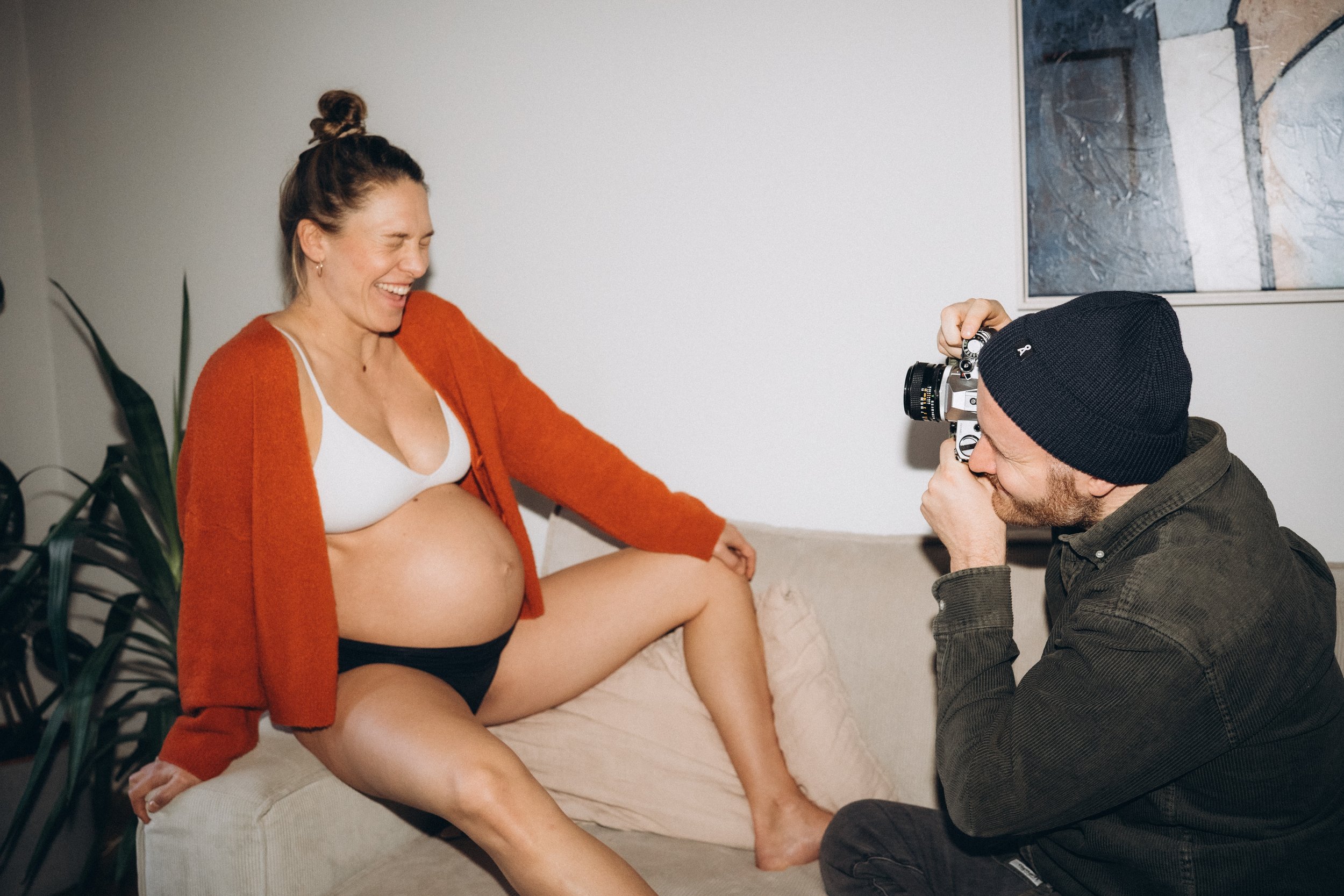 Eine schwangere Frau sitzt lachend auf einer Couch, hält ihr Bein hoch, während ein Fotograf sie mit einer Kamera fotografiert.