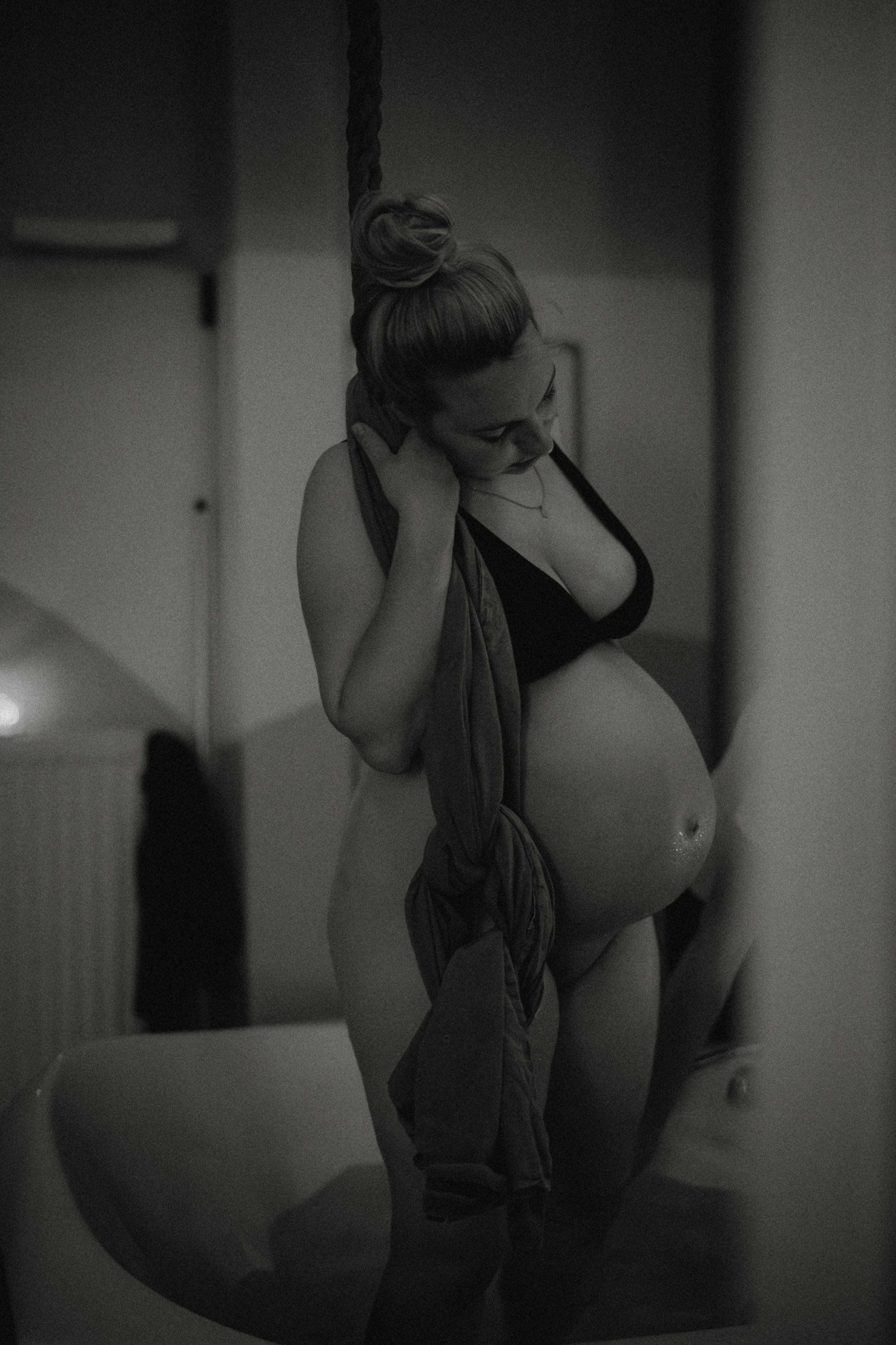 Schwangere Frau in Unterwäsche, die im Badezimmer steht, sich eine Jacke über die Schulter legt, in einem schwarzen und weißen Bild.