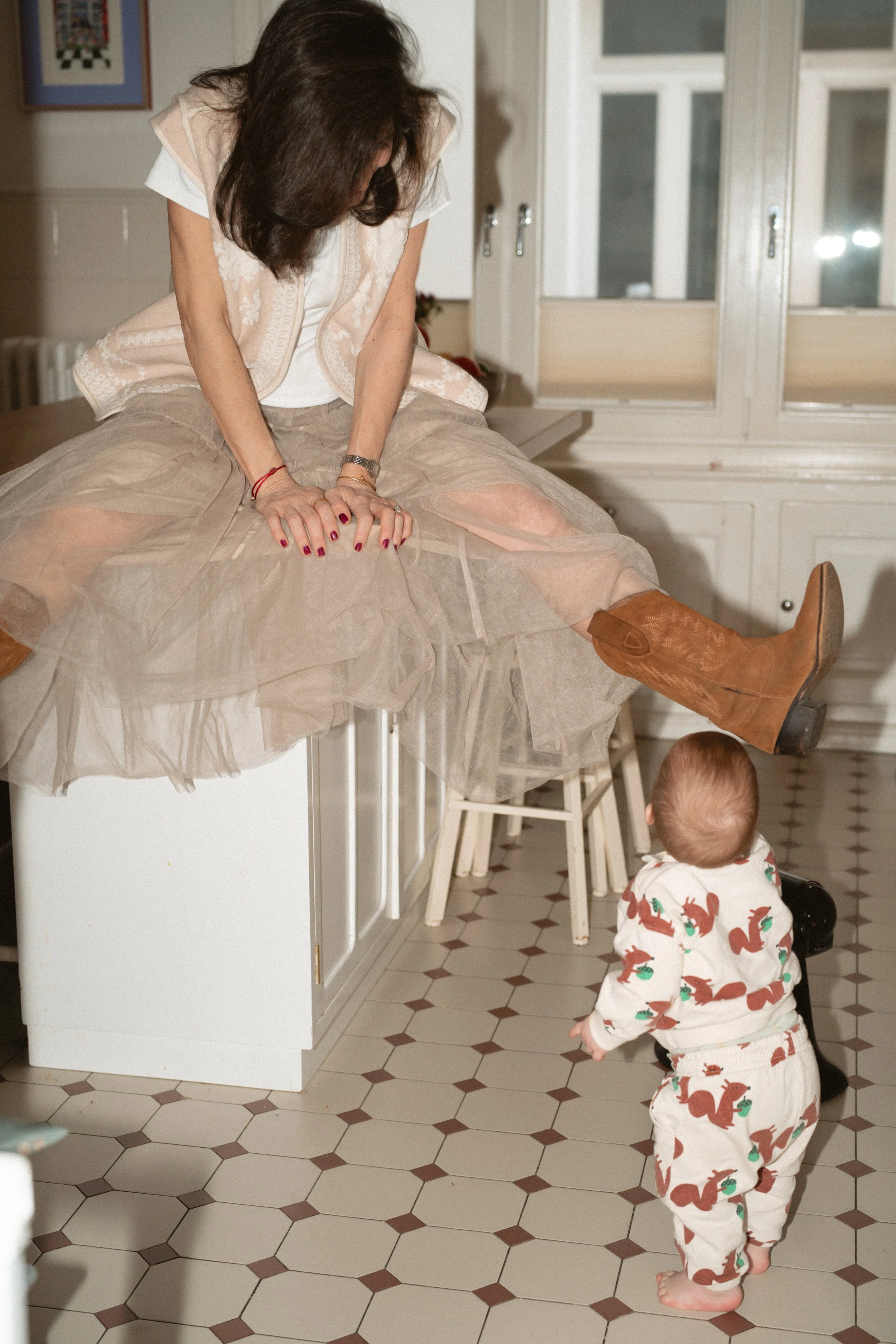 Eine Frau sitzt auf einer Küchenarbeitsplatte, während ein Baby mit einem Tiermusterpyjama vor ihr steht, im Küchenraum mit Fliesenboden.