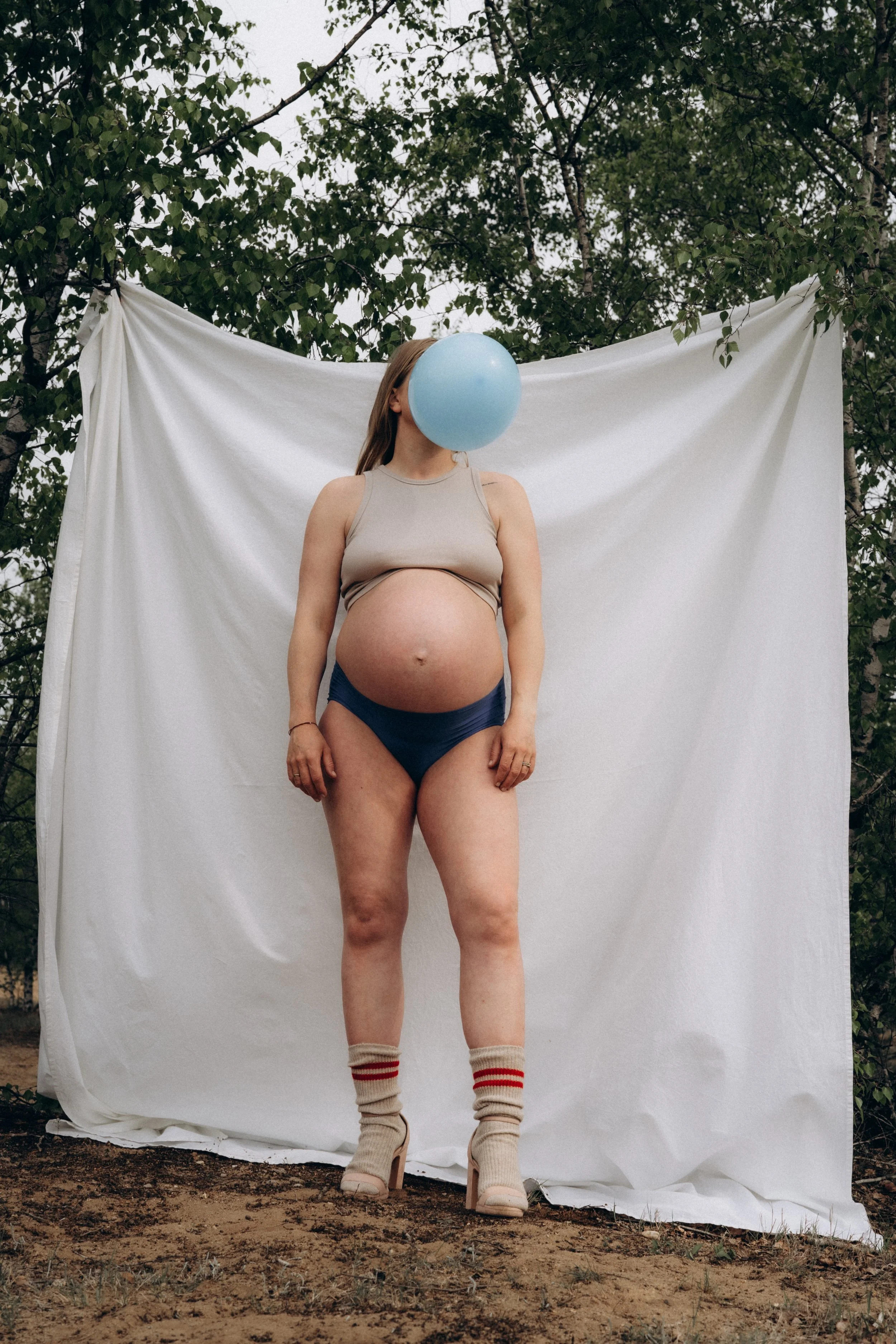 Eine schwangere Frau steht vor einem weißen Tuch im Freien, hält einen blauen Ballon vor ihr Gesicht und trägt hohe Schuhe sowie Socken.