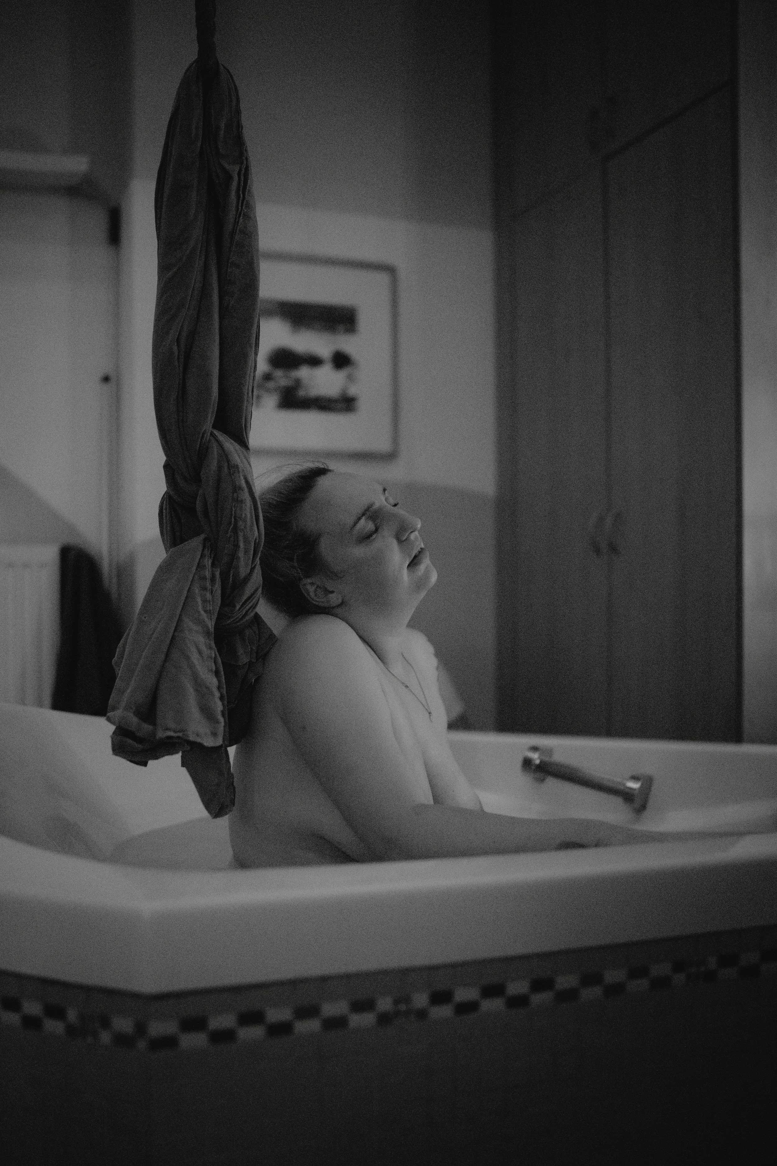 Schwarz-Weiß-Foto einer Frau in einer Badewanne, entspannt mit geschlossenen Augen, Hand auf dem Wasser, in einem Badezimmer mit Kleidung auf einem Haken und einem Schrank im Hintergrund.