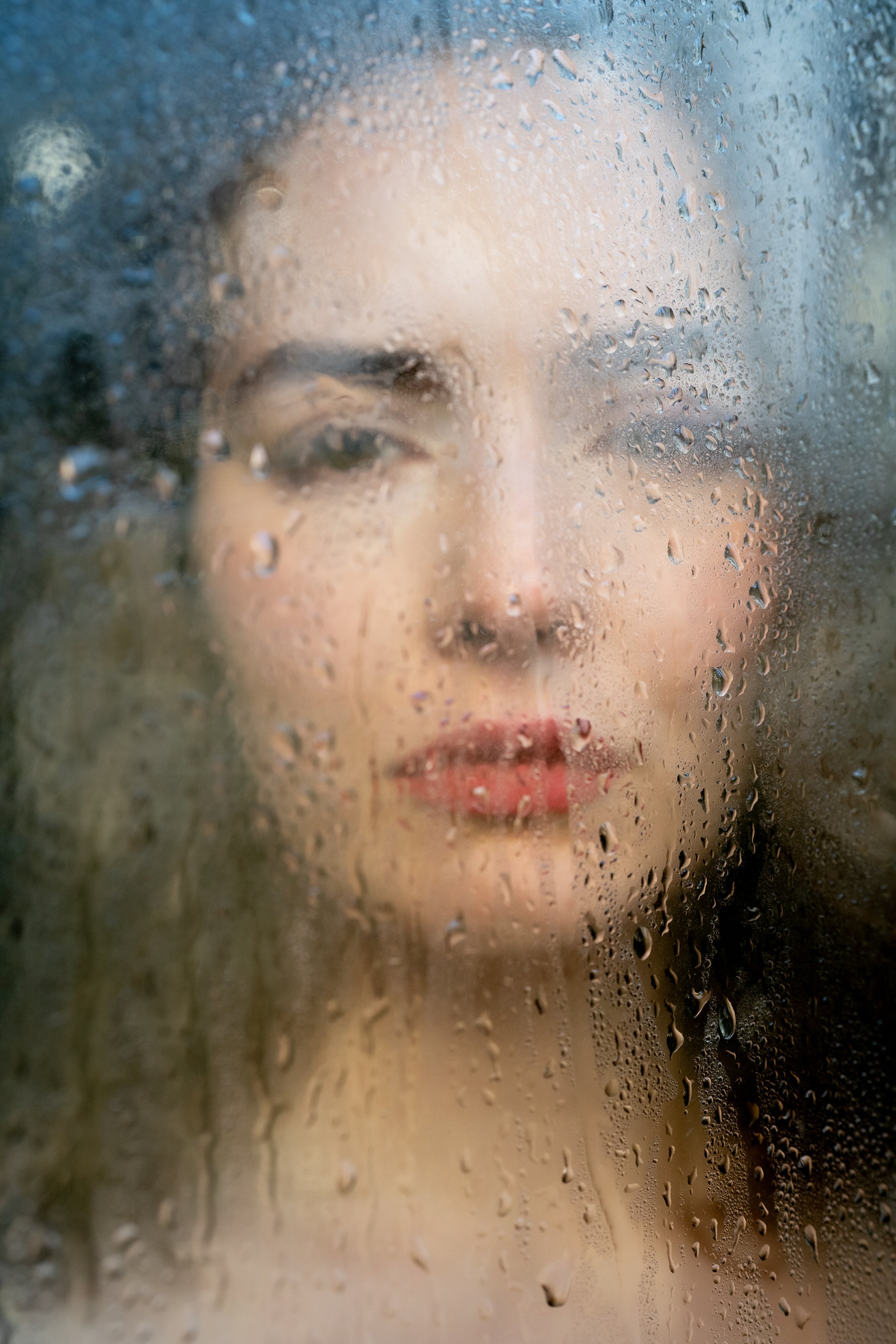 Nahaufnahme eines Gesichts einer Frau, sichtbar durch einen beschlagenen Fenster mit Regentropfen.