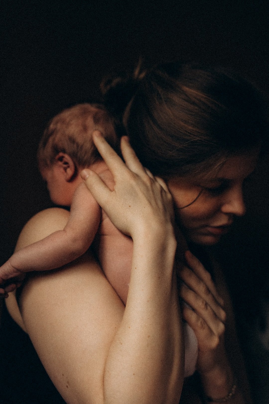 Eine Frau hält ein Baby liebevoll an ihrem Hals, beide sind nackt und vor einem schwarzen Hintergrund. Die Frau schaut nach unten, das Baby hat den Kopf an ihrer Schulter.