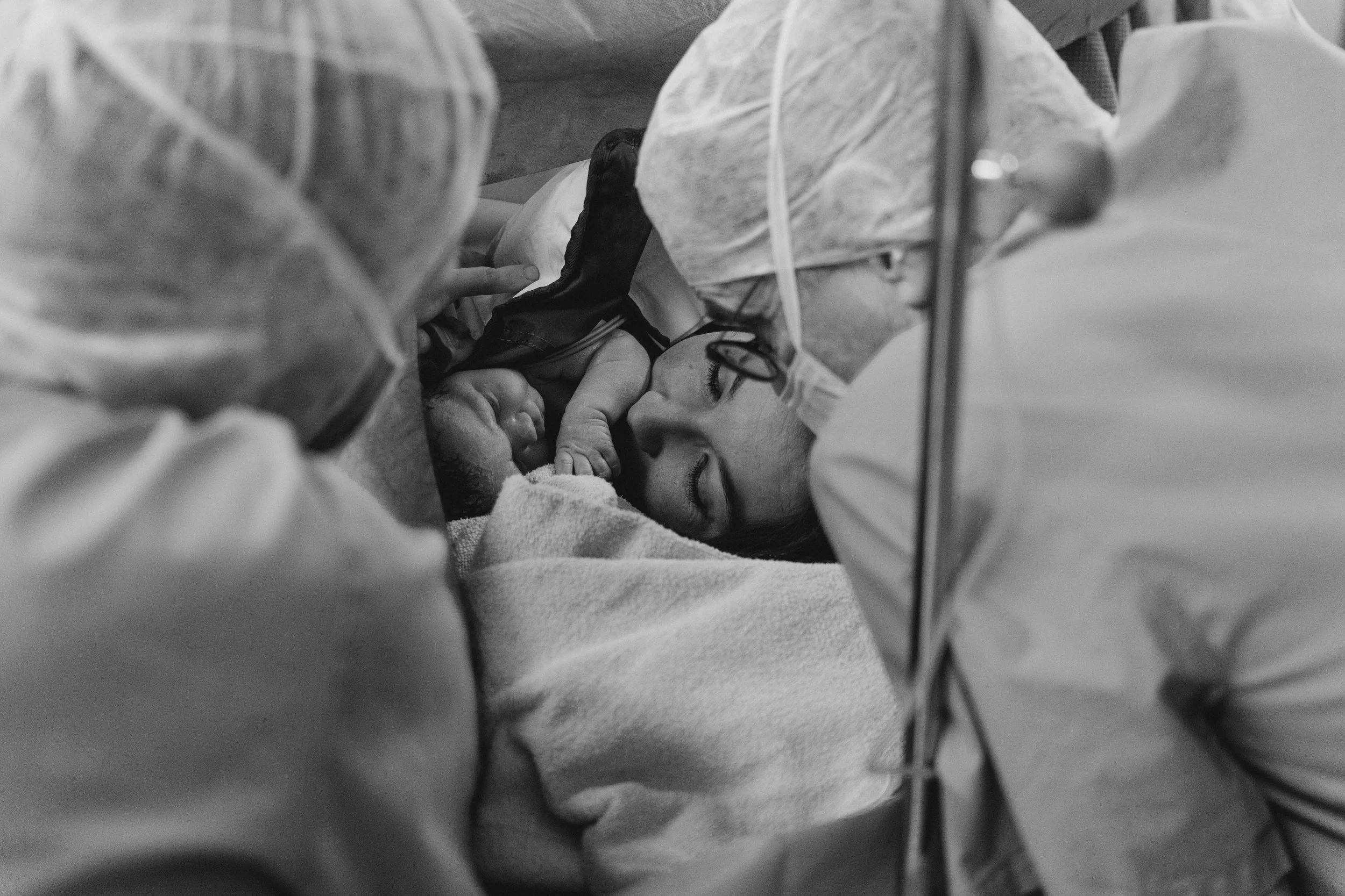 Eine Frau liegt in einem Krankenhausbett, sichtbar durch Schutzkleidung und eine OP-Haube, während sie ein Neugeborenes hält, das offenbar gerade geboren wurde. Die Szene ist in Schwarz-Weiß dargestellt.