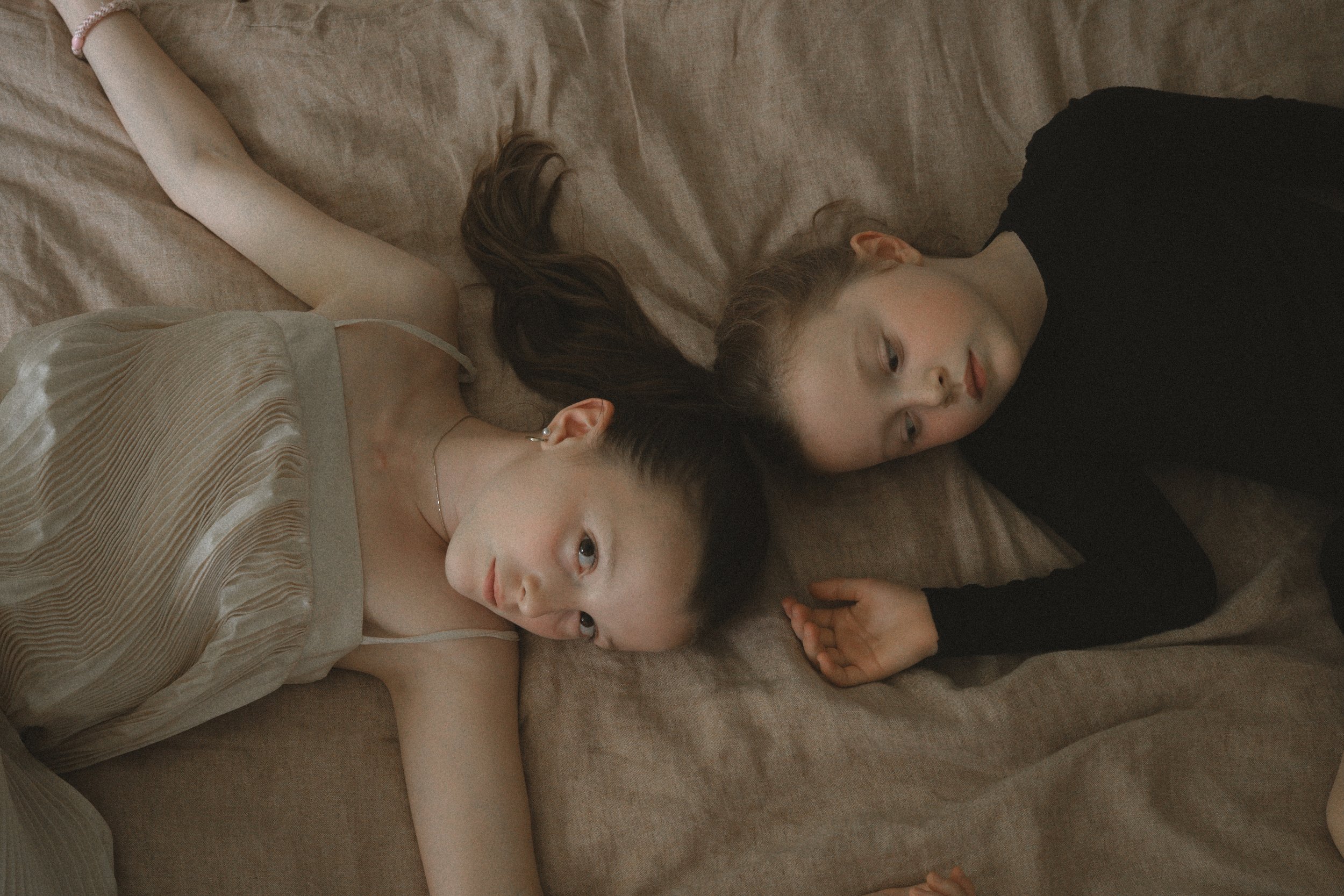 Zwei junge Mädchen liegen auf einem Bett, Blick nach oben gerichtet, entspannte Stimmung, eines trägt ein cremefarbenes Kleid, das andere ein schwarzes Oberteil.