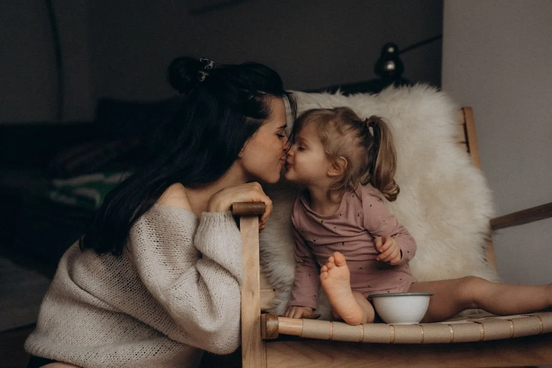 Eine Frau und ein kleines Mädchen, die sich küssen, sitzen auf einem Holzstuhl in einem dunklen Raum.