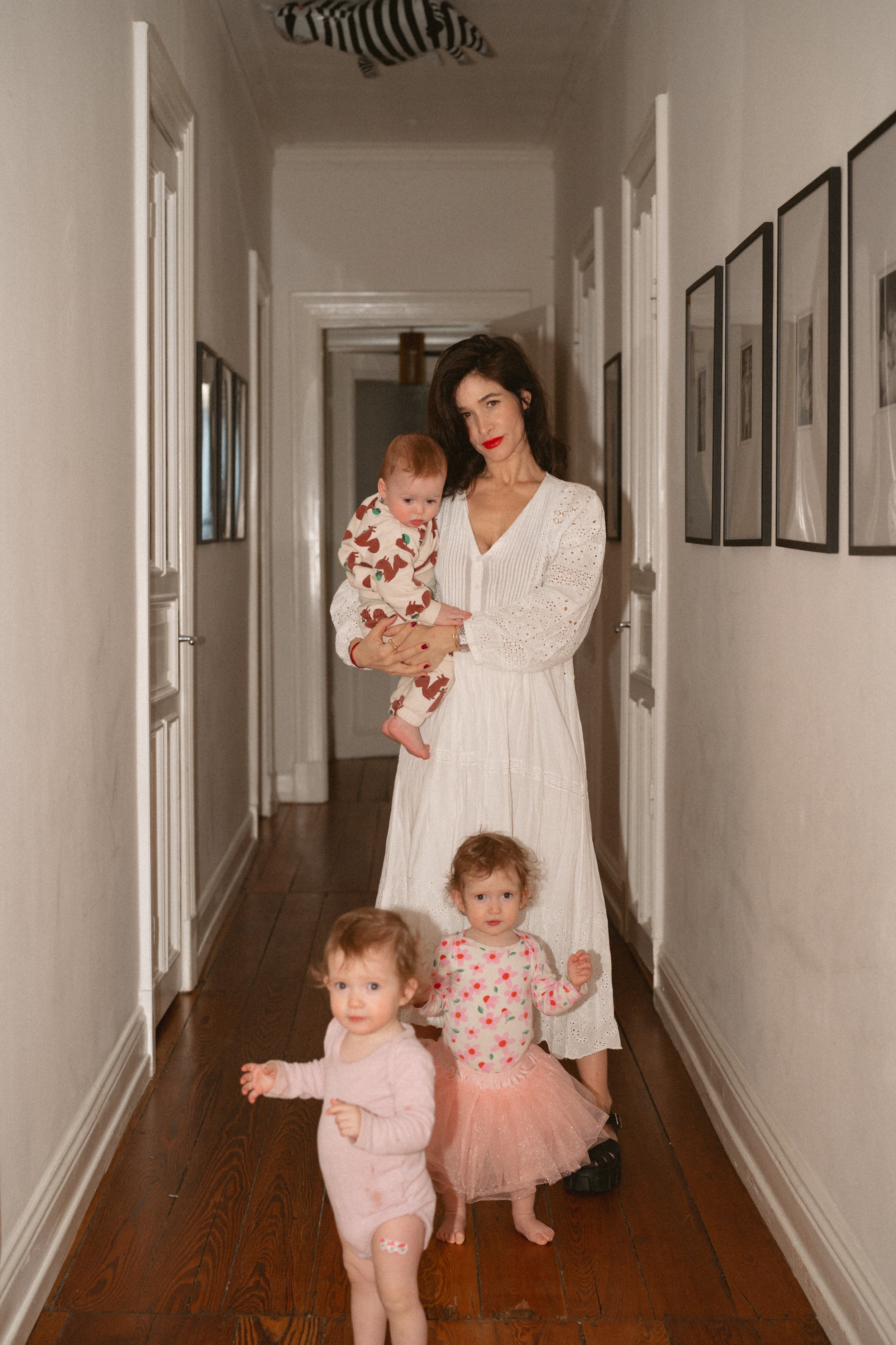 Eine Frau mit dunklen Haaren in einem weißen Kleid steht in einem Flur, die Arme um drei kleine Kinder. Die Kinder tragen bunte Kleidung, ein Mädchen hat einen Tüllrock. Im Hintergrund sind Wandbilder und eine zebra-Form auf der Decke sichtbar.