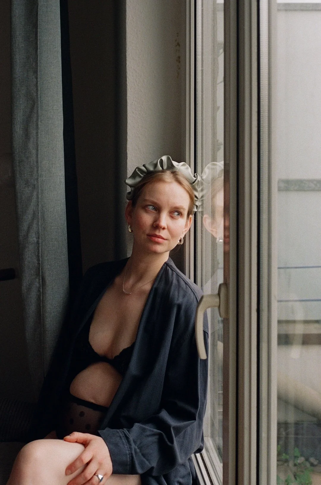 Eine junge Frau mit rotem Haar sitzt am Fenster, trägt ein schwarzes Outfit und einen Haarschmuck, blickt nach draußen.