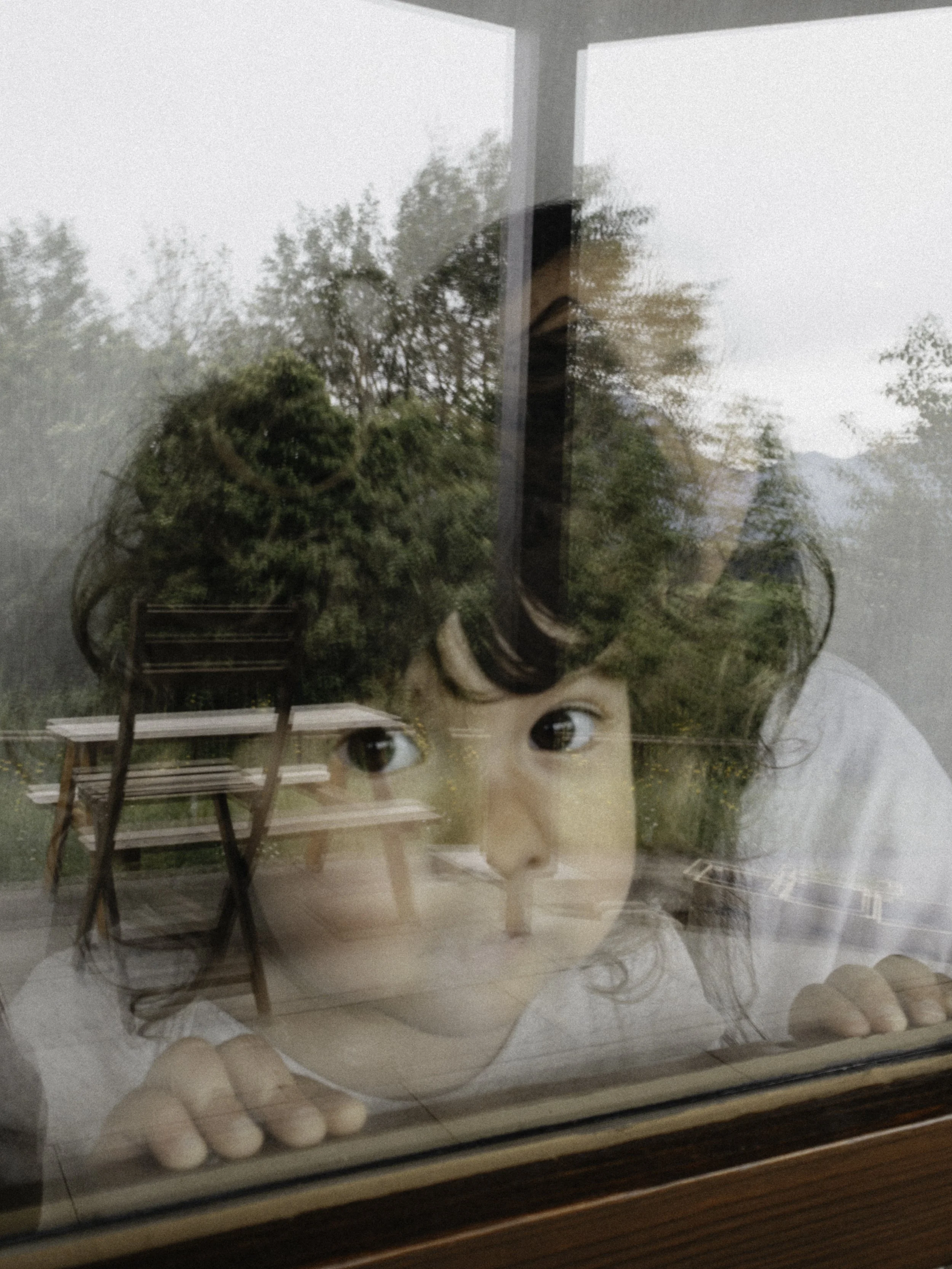 Ein junges Mädchen schaut durch eine Fensterscheibe, die die Reflexion eines Außenbereichs mit Bänken und Bäumen zeigt.