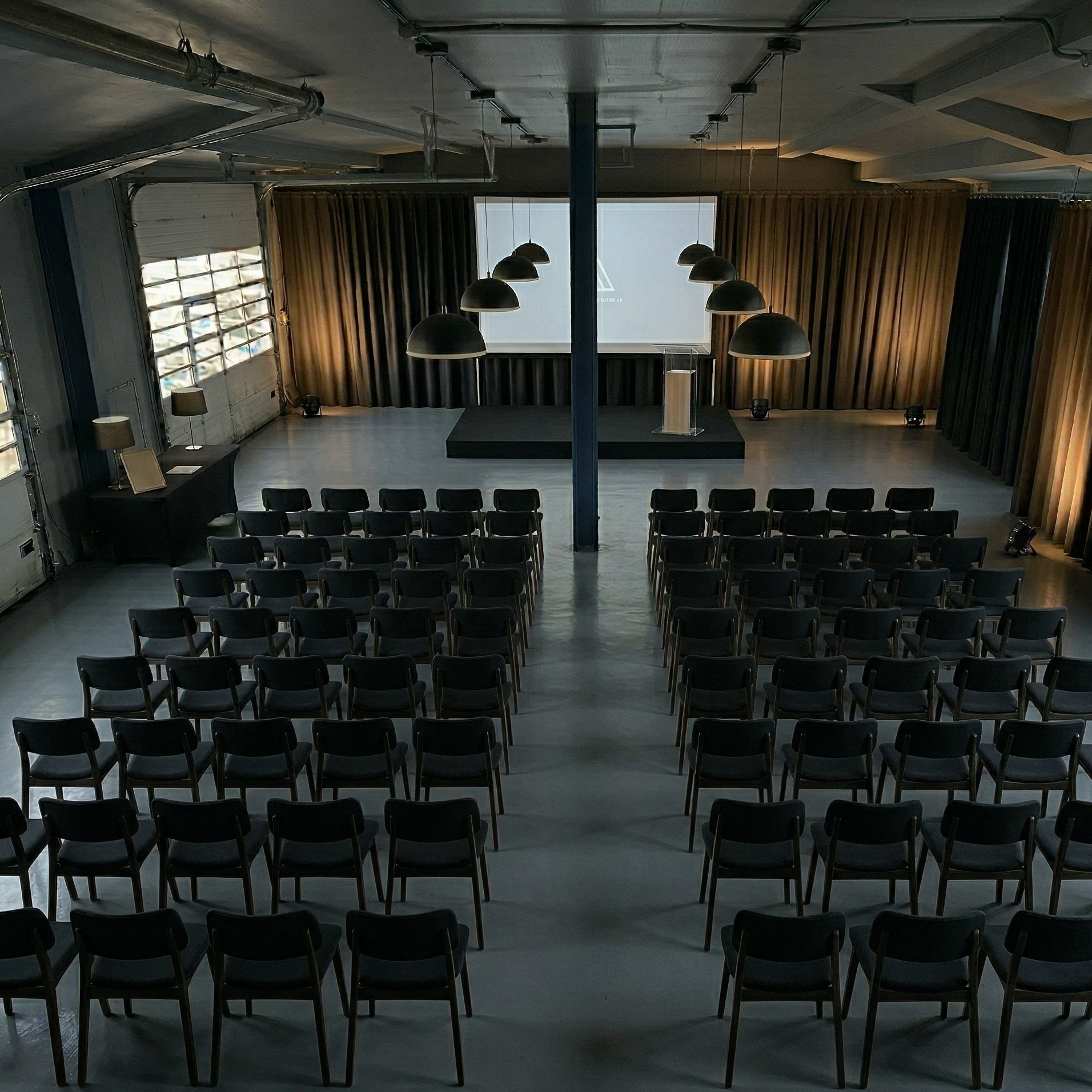 Empty conference room with rows of black chairs facing a stage with a podium and a large projection screen. The room has curtains and hanging lights.