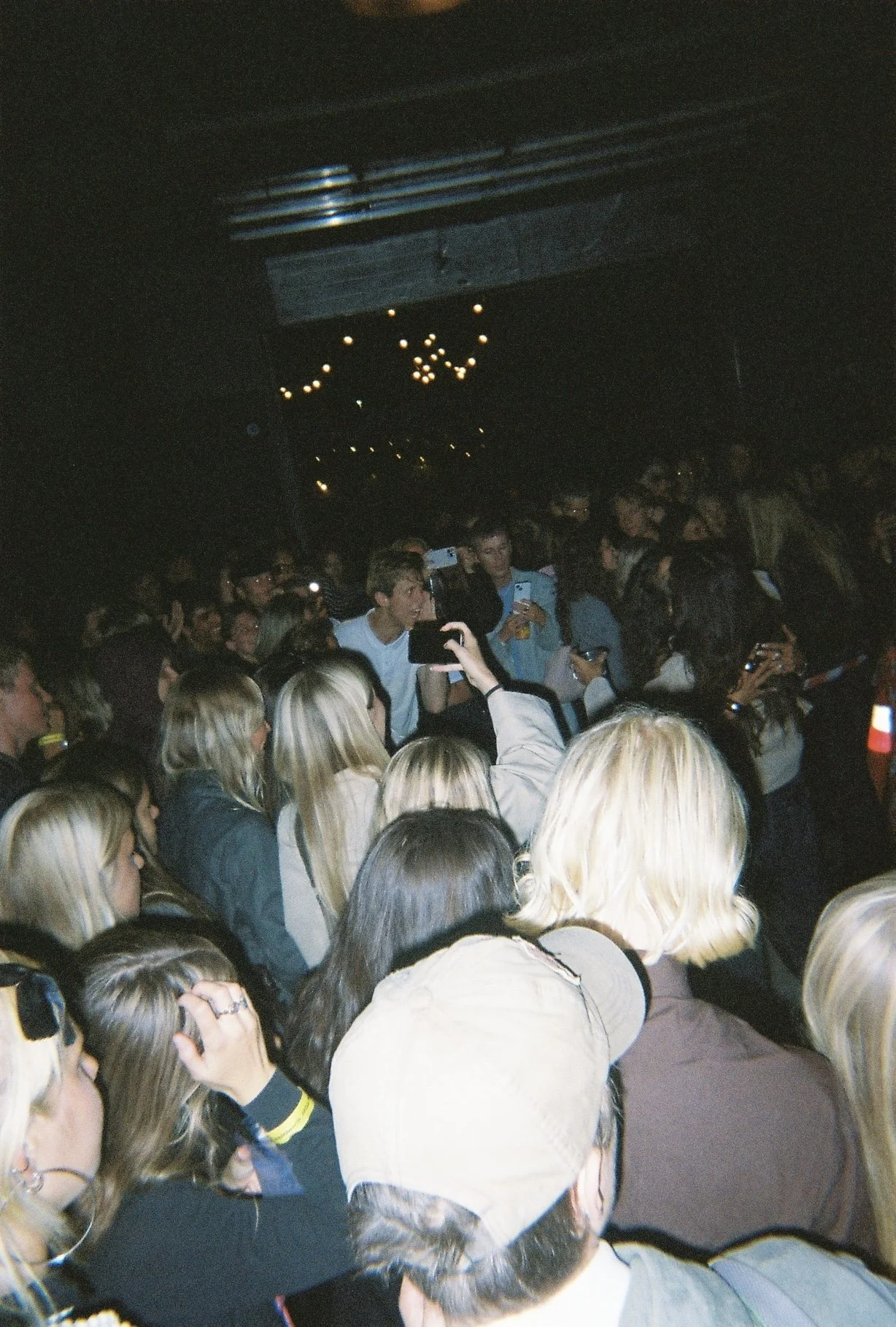 Guests gathered under string lights at an indoor evening party, with several people holding up phones.