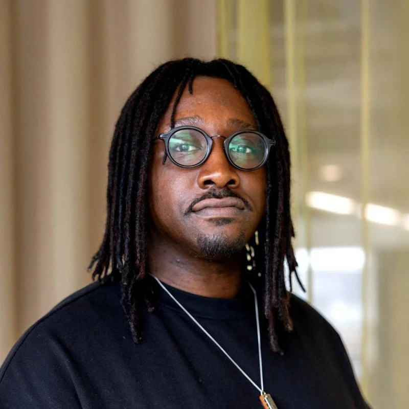 Portrait of a man with dreadlocks and glasses, wearing a black shirt, standing indoors near a window with blinds.
