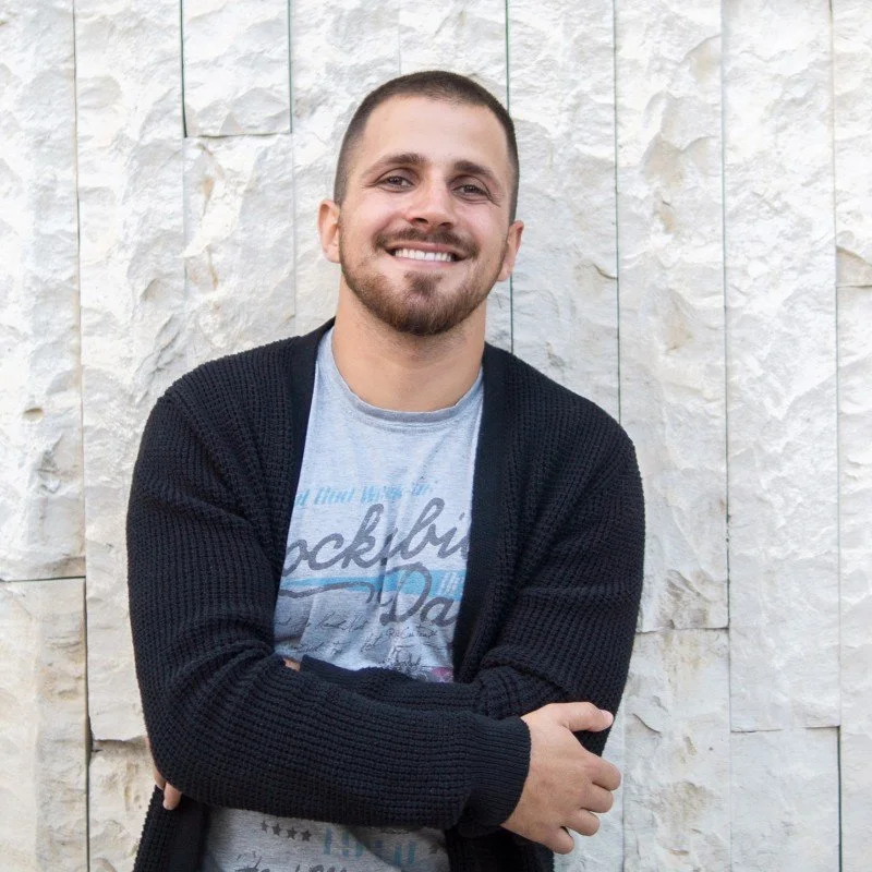 A young man with a beard and short hair smiling, standing with arms crossed in front of a textured light-colored stone wall, wearing a light gray graphic t-shirt and a black knitted cardigan.