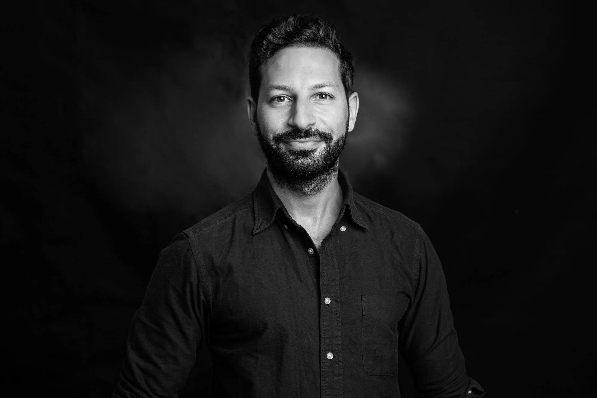 Black and white portrait of a man with a beard and short hair, wearing a dark button-up shirt, smiling slightly, against a dark background.