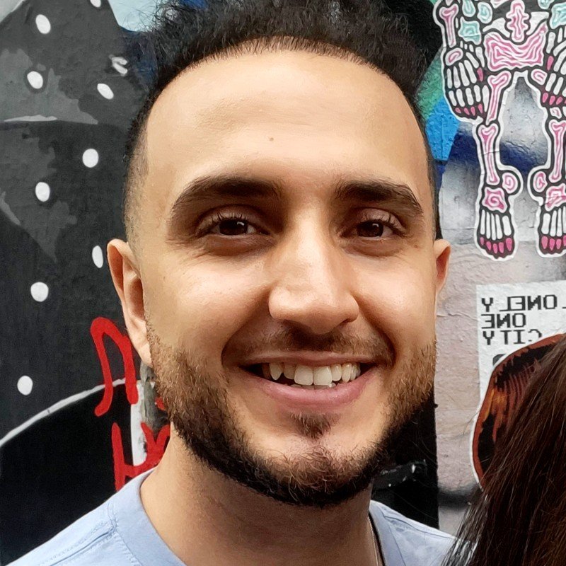 Close-up of a smiling young man with short dark hair and a beard, standing in front of a colorful graffiti mural.