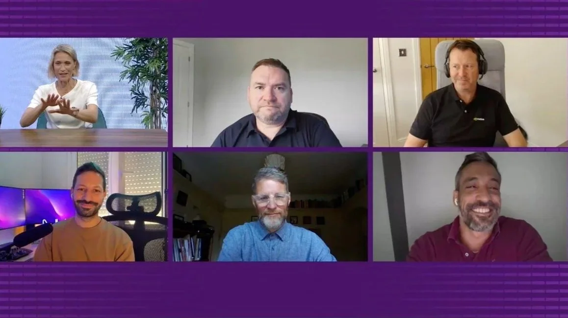 Six people participating in a virtual meeting, each in their own video frame. The top left woman has gray hair and is gesturing with her hands, the top middle man has short dark hair and a serious expression, the top right man has headphones and is sitting in a beige chair. The bottom left man has a beard and is sitting in front of a multi-monitor setup, the bottom middle man has glasses and a beard, and the bottom right man is smiling and has a beard.