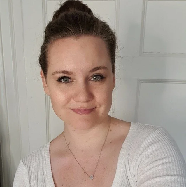 A young woman with brown hair tied up in a bun, wearing a white ribbed top and a delicate necklace, smiling in front of a white door.