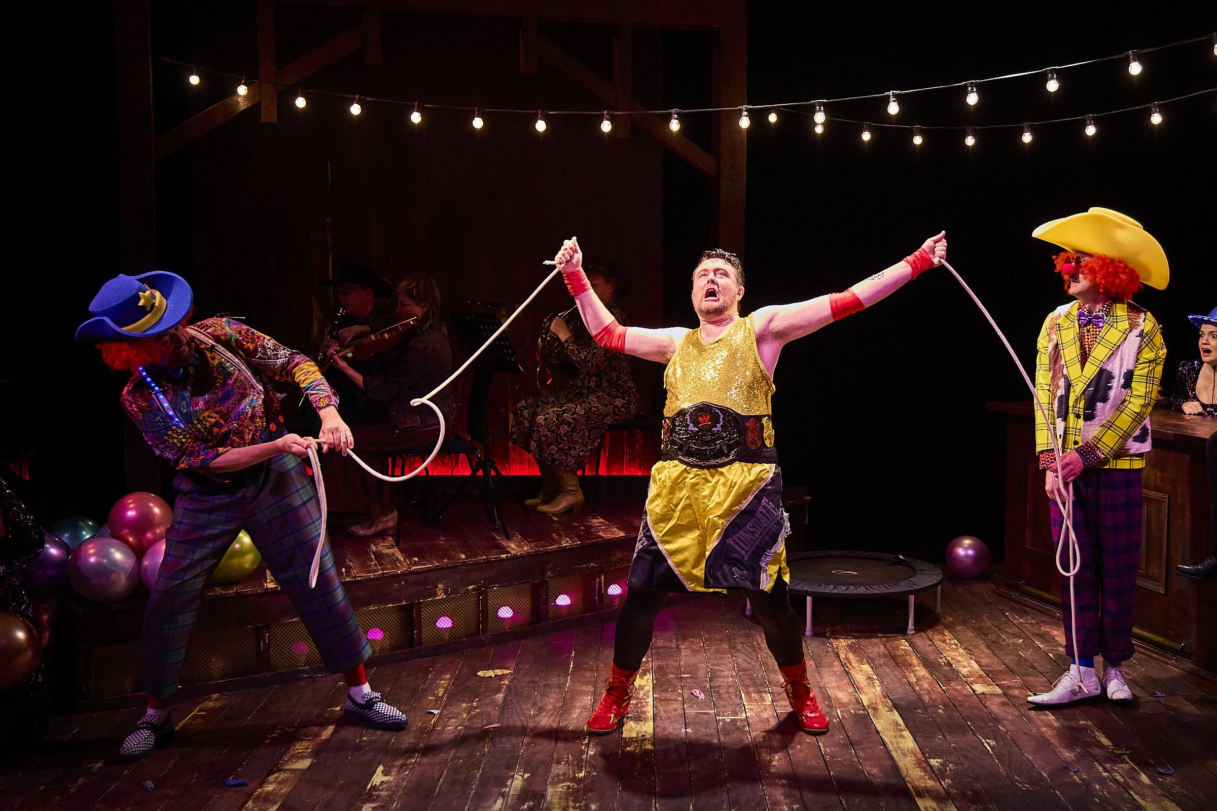 A group of performers dressed as clowns, including one in a yellow vest and red shoes, performing on stage with balloons and balloons on the floor, with string lights hanging above them.