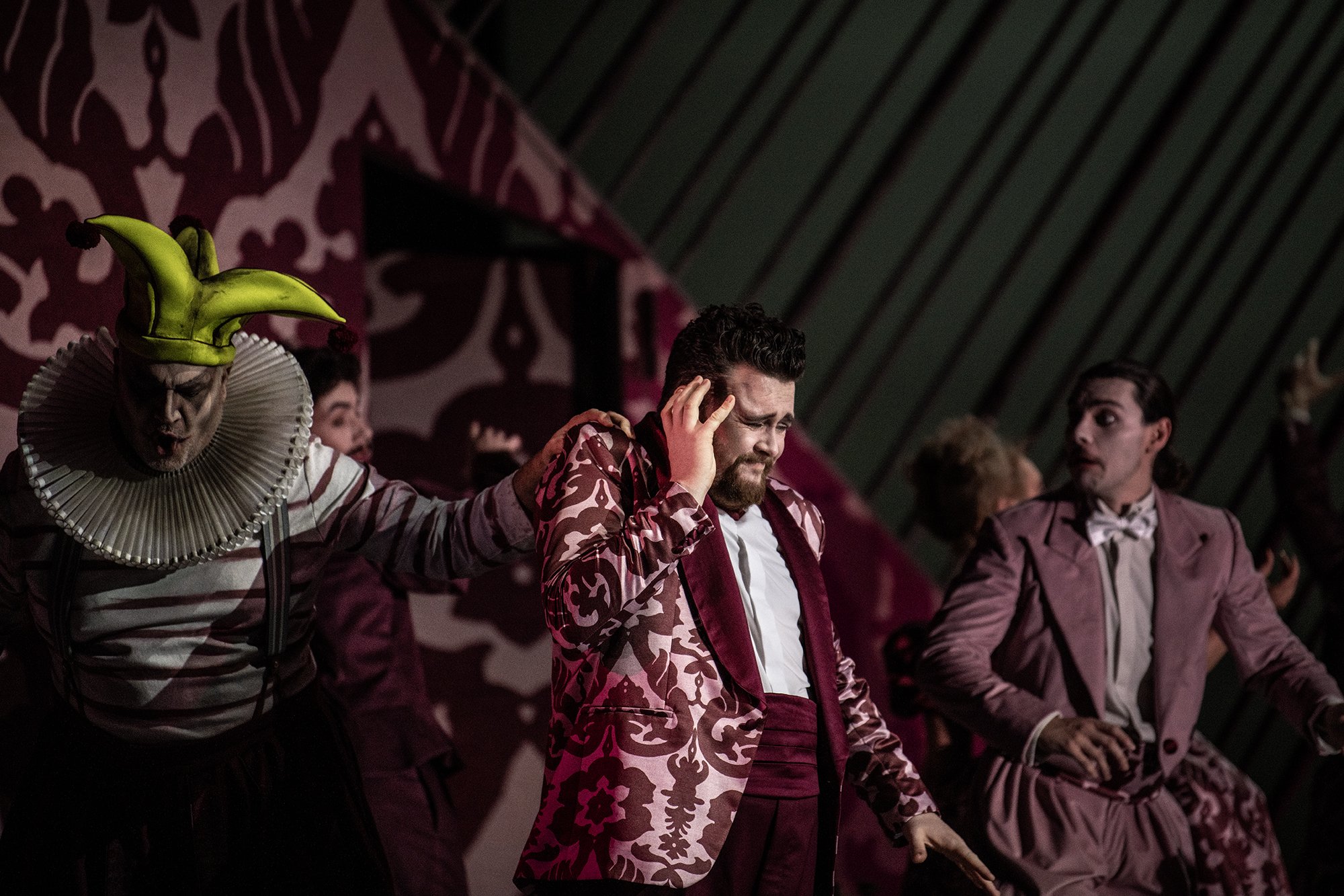 People dressed in stylish, patterned suits on stage, with one person wearing a balloon hat. They appear to be performing or at an event, with a patterned backdrop and dim stage lighting.