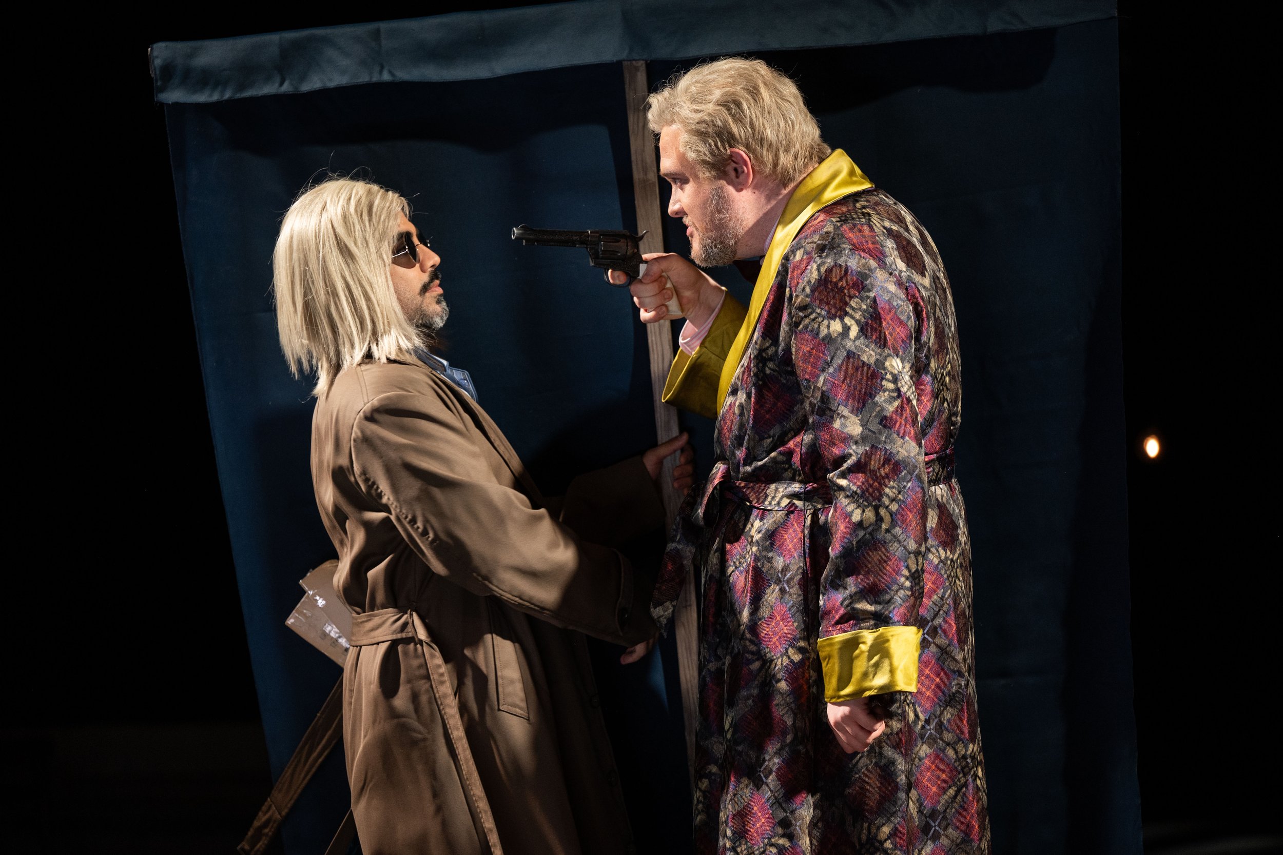 Two men with blonde hair facing each other, one holding a gun pointed at the other's head, with a dark background and a blue curtain.
