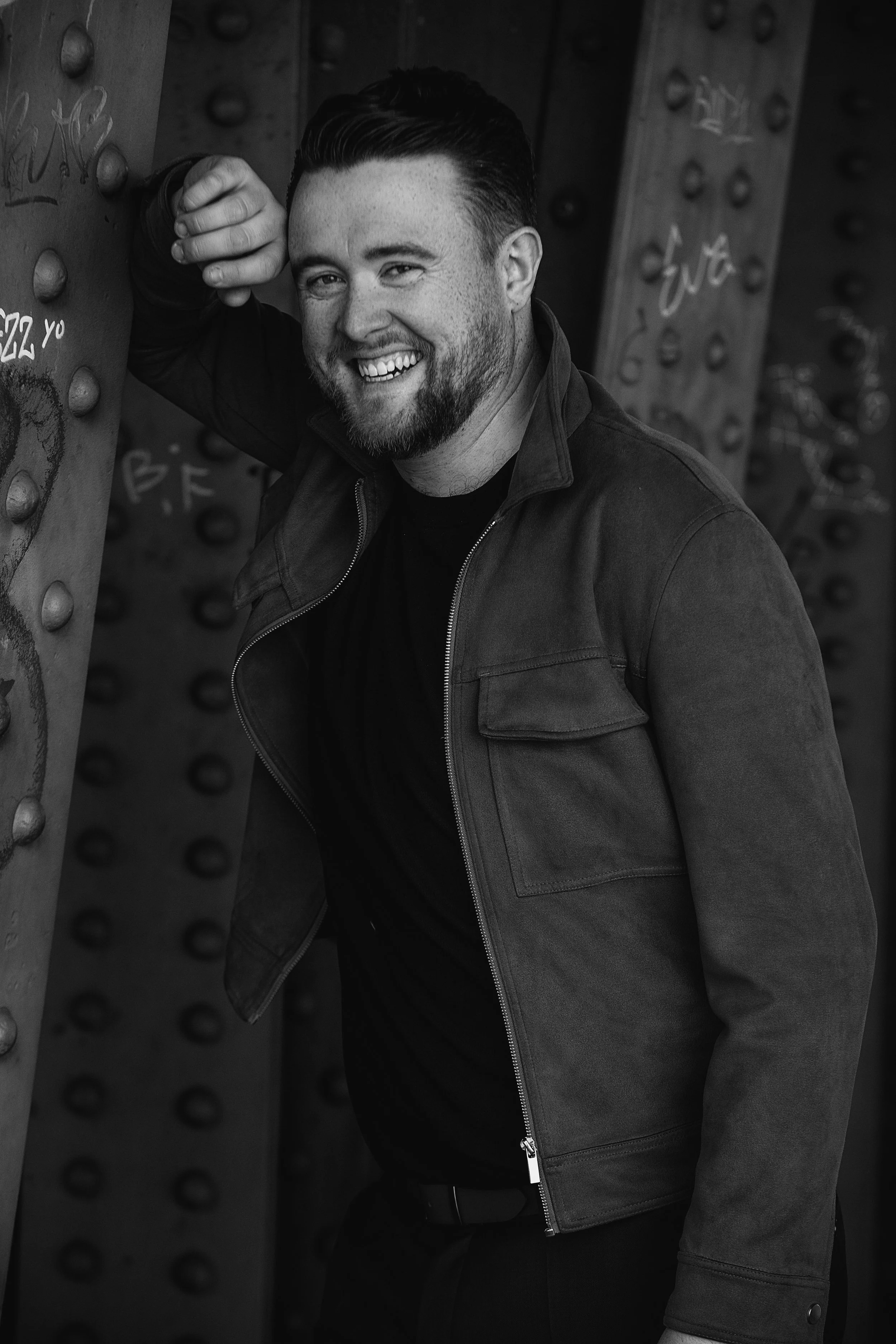 A smiling man with dark hair and a beard, wearing a dark jacket, leaning against a graffiti-covered metal structure with rivets, in an urban setting.
