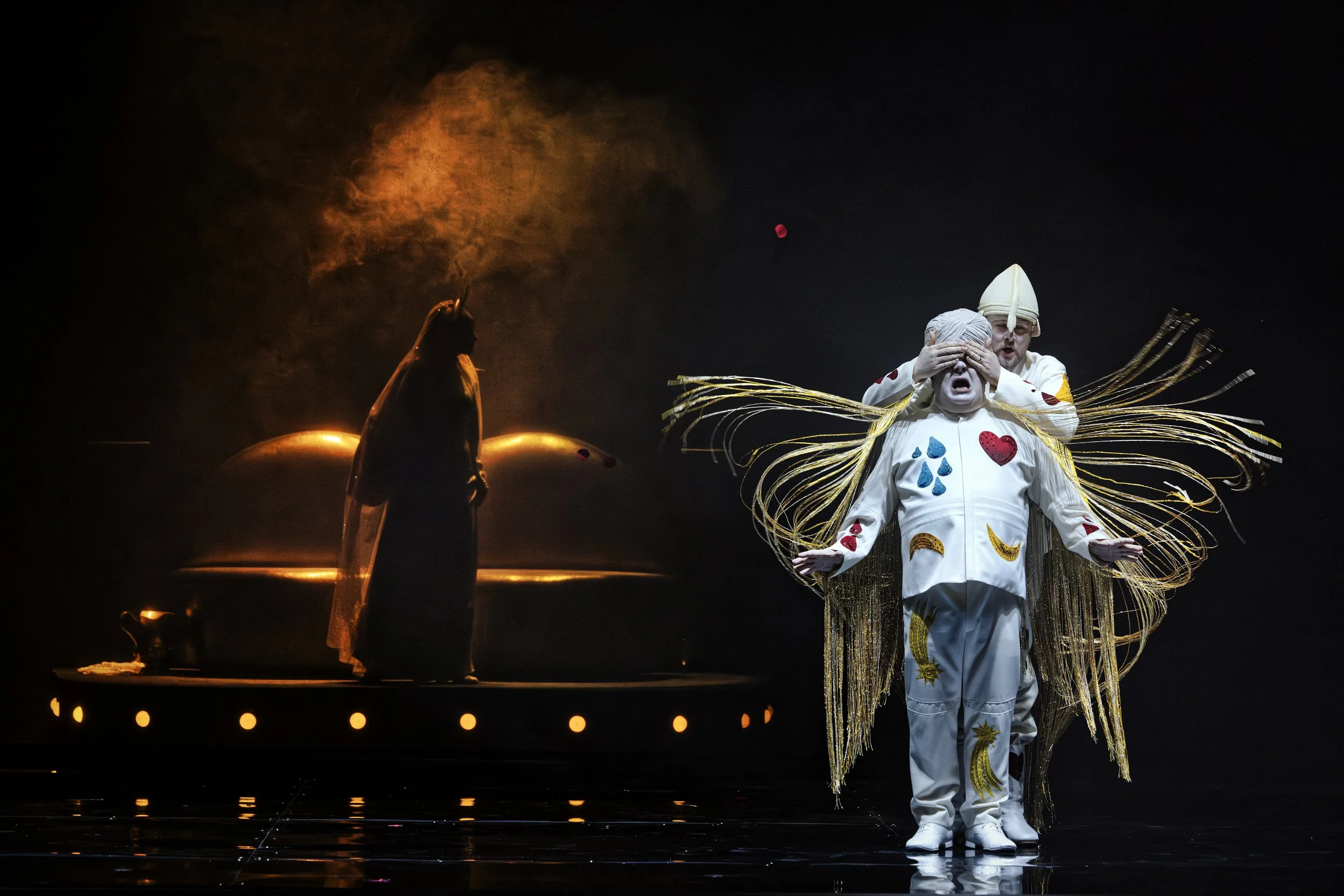 A theatrical performance with people dressed in colorful costumes, including a performer with angel-like wings and a hat, standing near water with flames and smoke in the background during a dark stage.