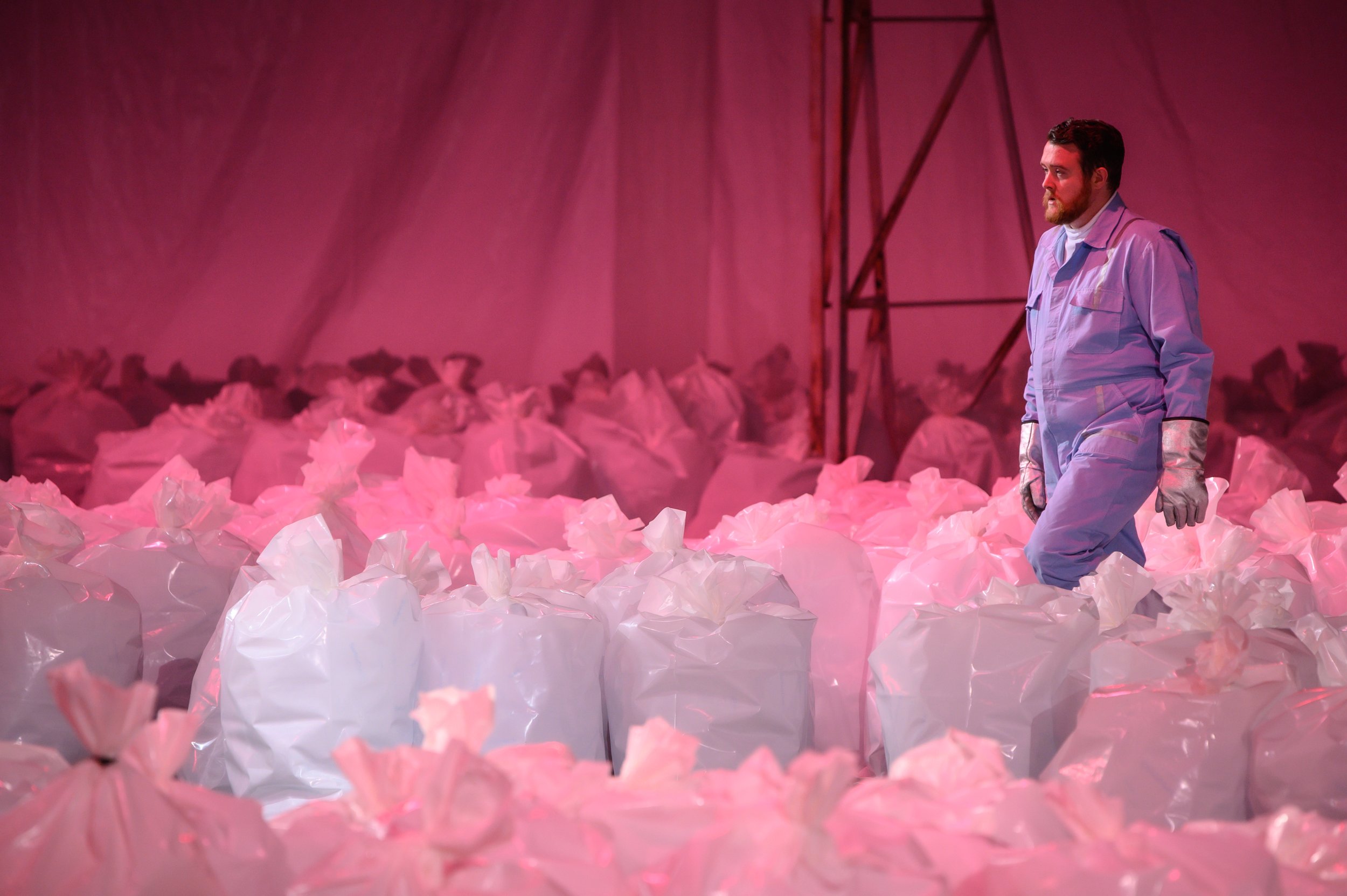 A man dressed in a white protective suit with gloves stands amidst pink and white crumpled plastic bags, with a pink backdrop and metal structure in the background.