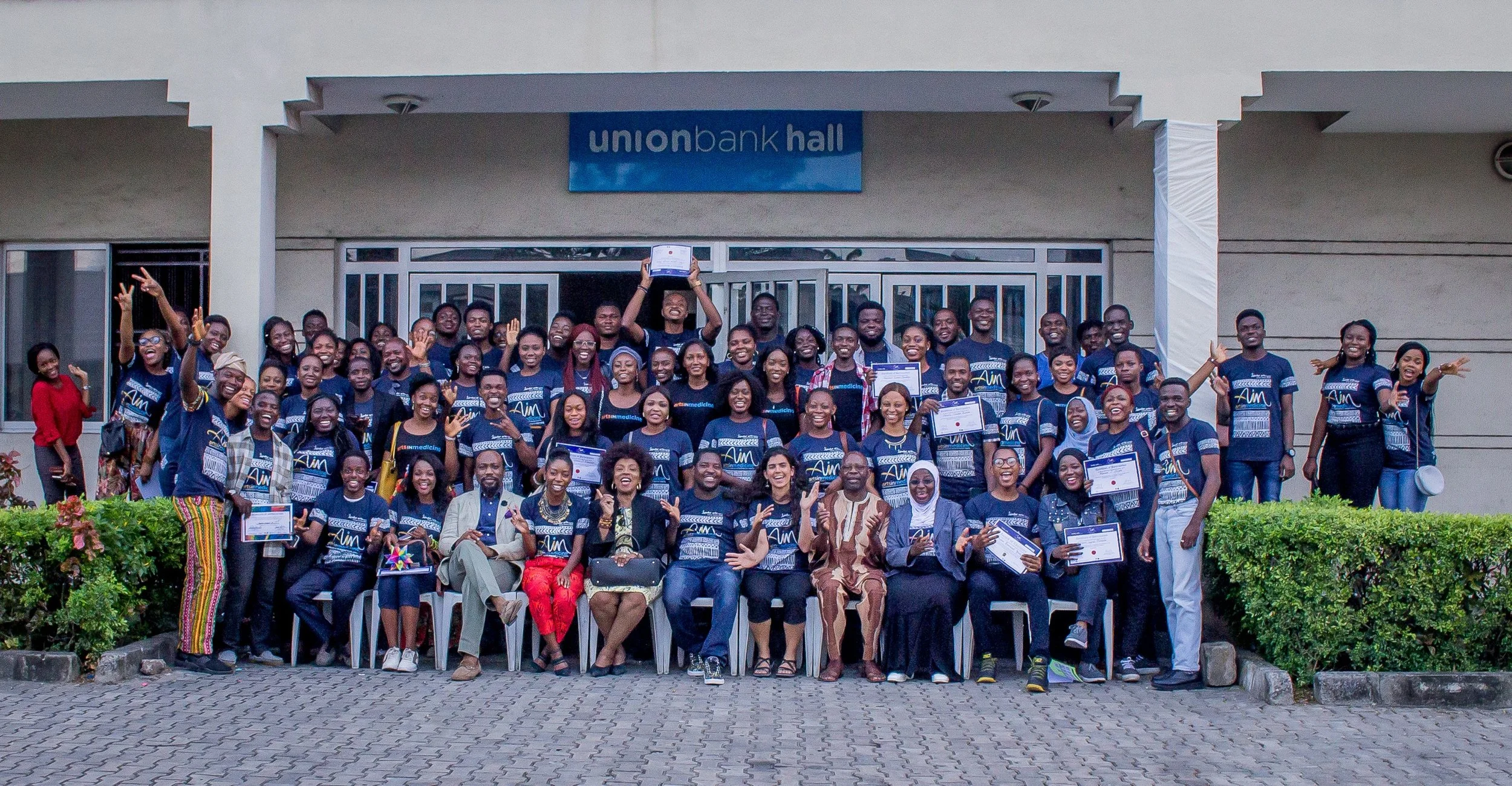 A large gathering of people in front of a building smiling jubilantly in navy blue shirts 