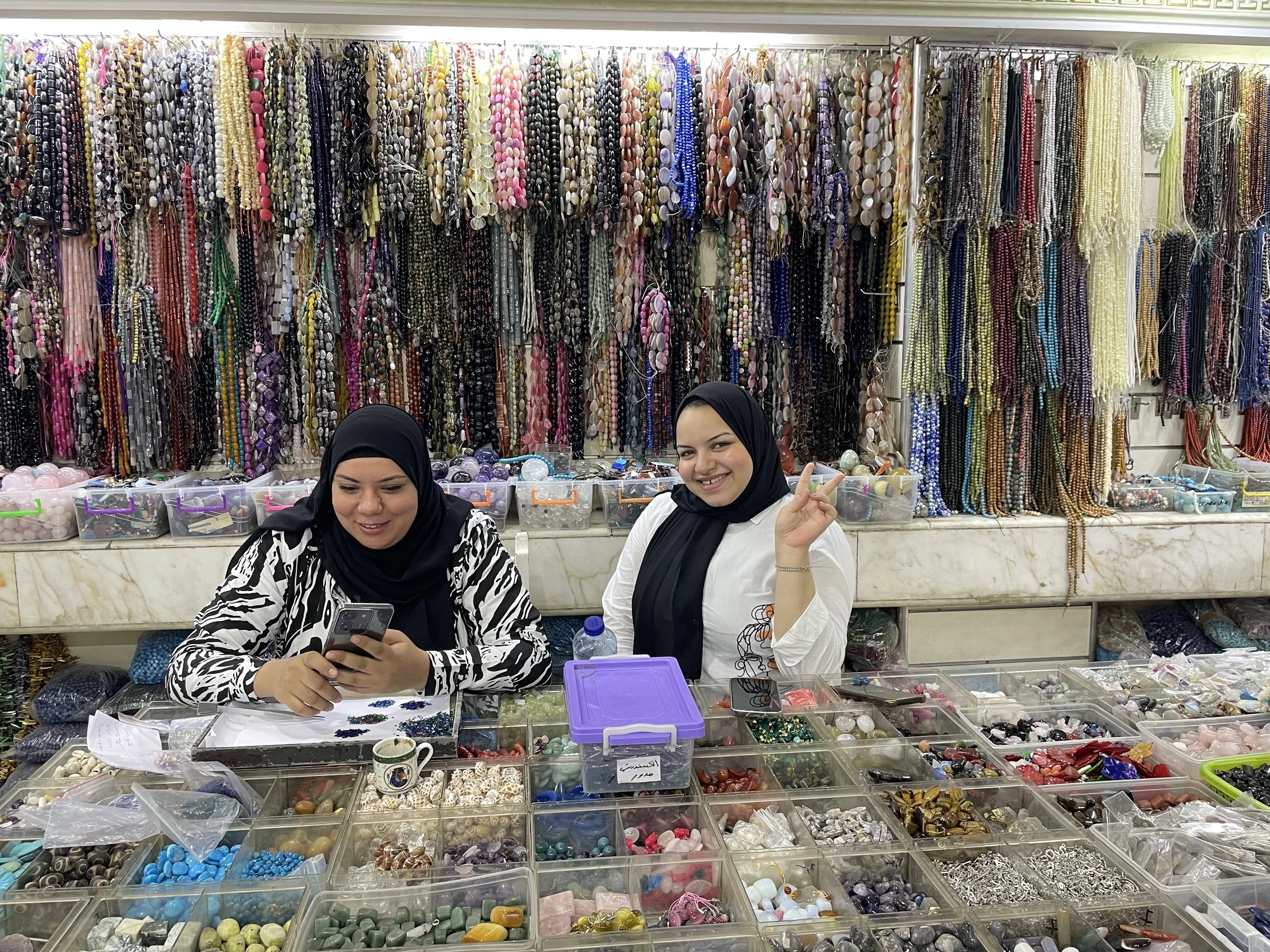 Two women smile while working at a store overflowing with beads and gems 