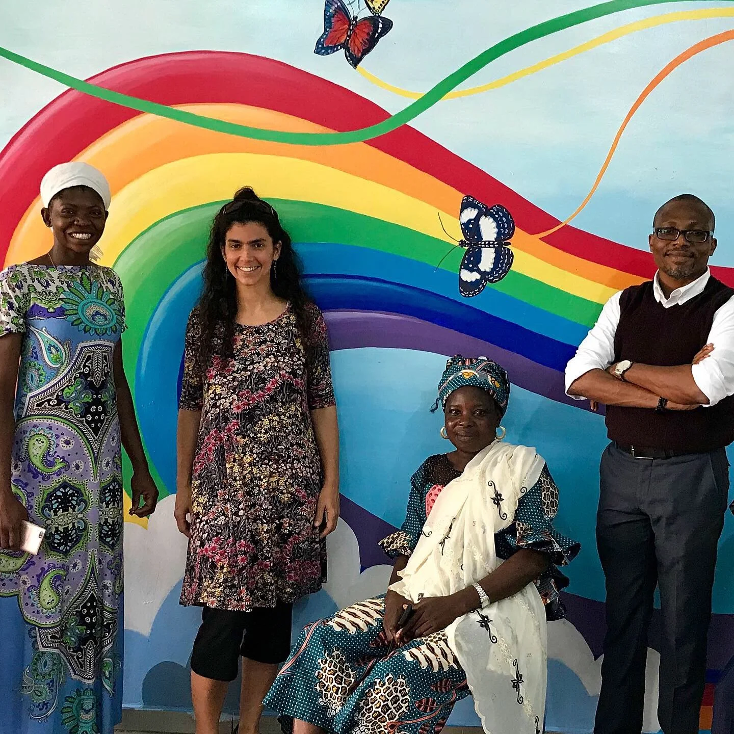 A group of proud people in colorful clothing stand in front of a mural 