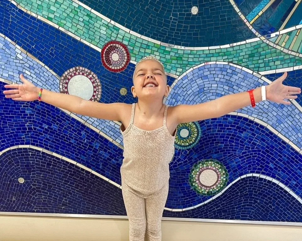 Young girl fighting cancer stands in front of mosaic mural 