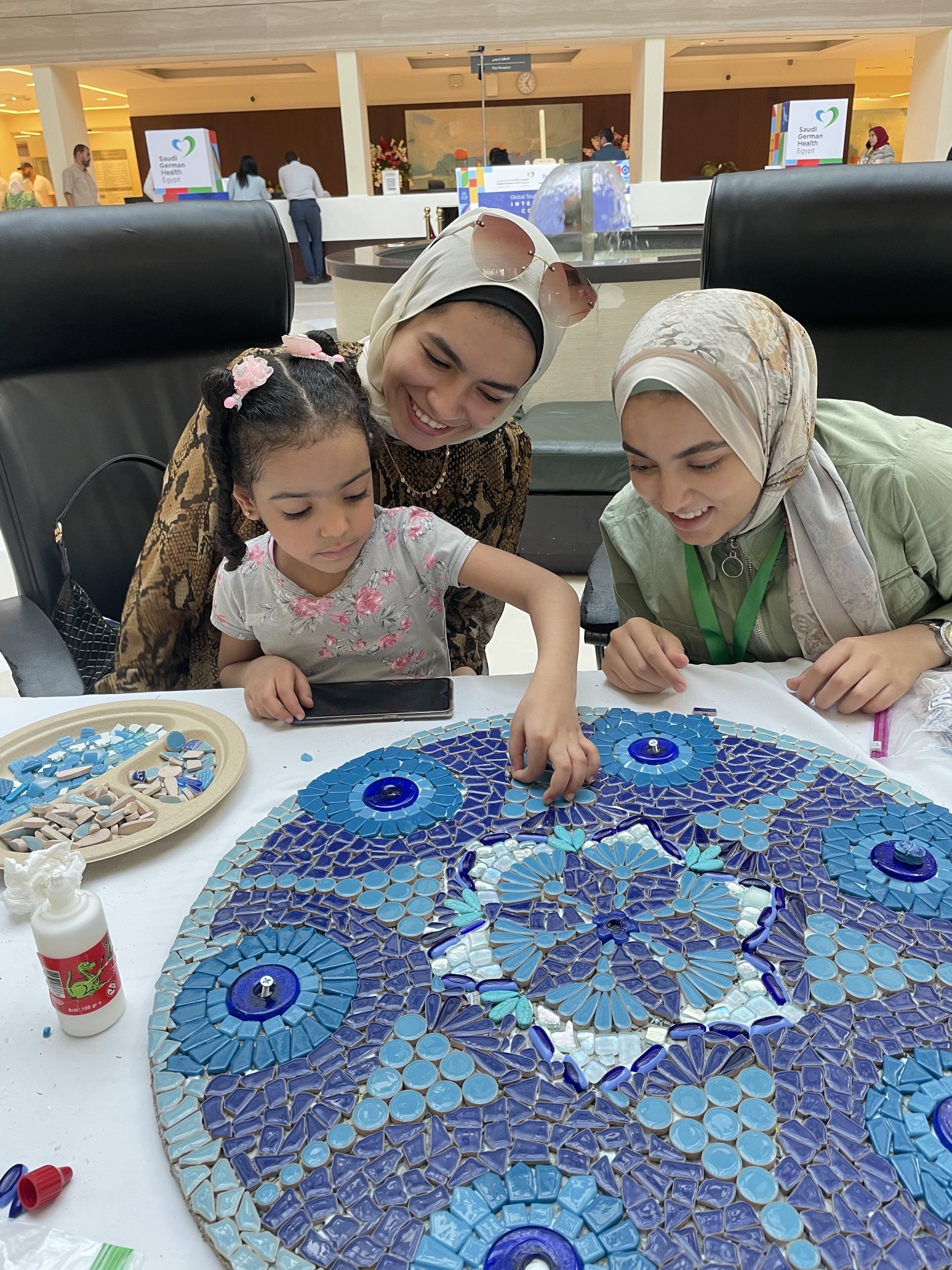 Two women and a girl work on a mosaic circle with blue tiles 