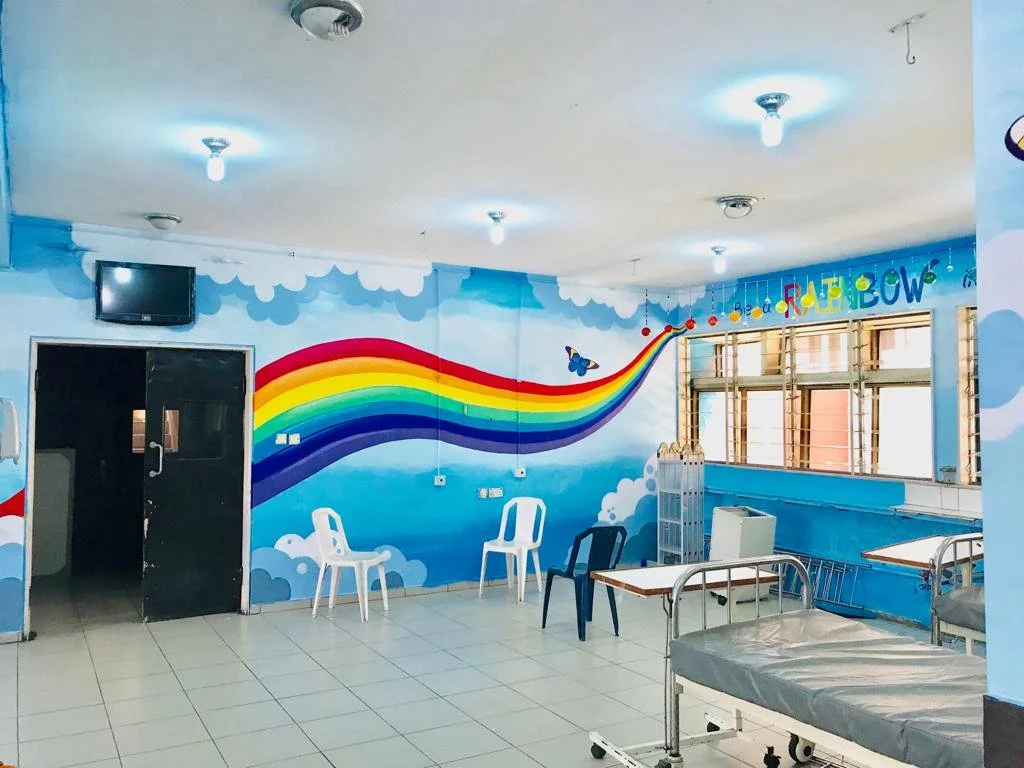 Plastic chairs and a hospital bed in front of a bright rainbow mural 