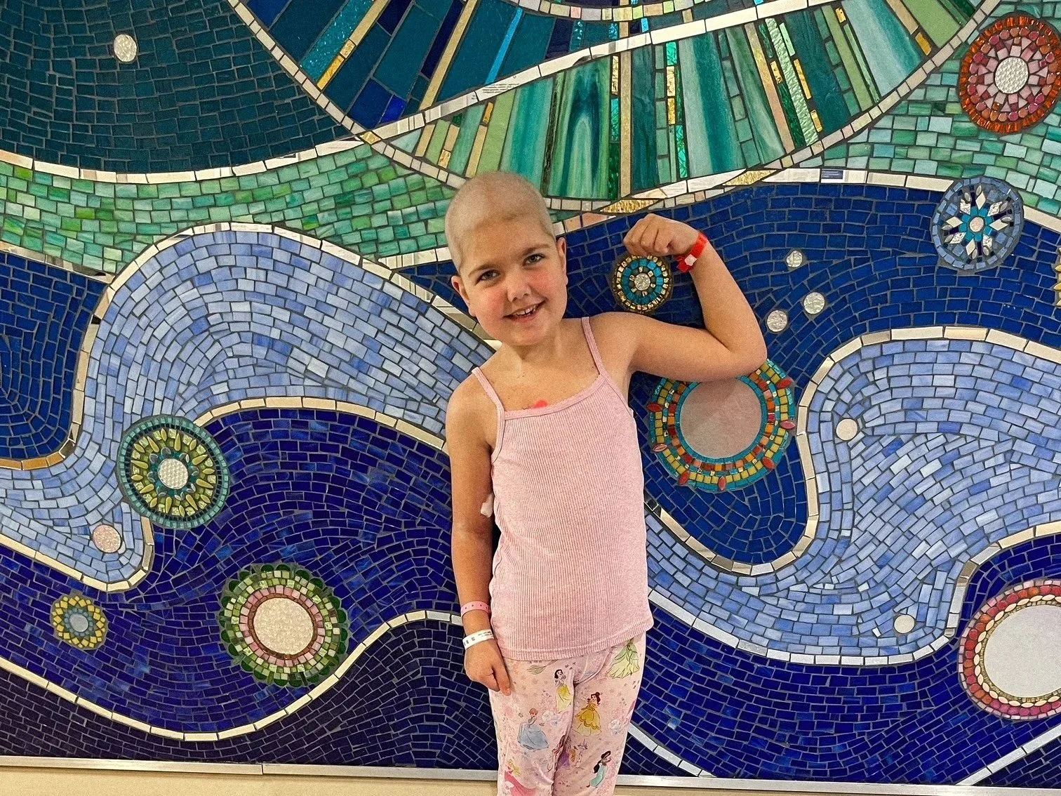 Young girl fighting cancer stands in front of mosaic mural 