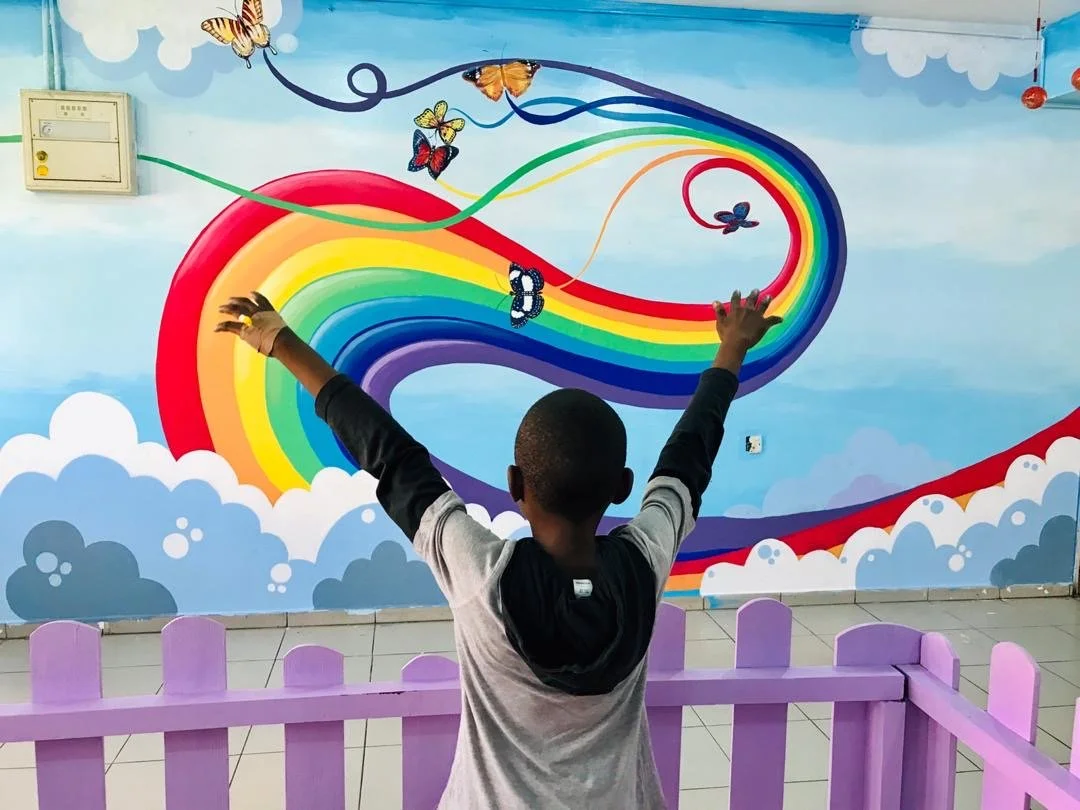 little boy reaches his hands upwards toward a mural of a flowing rainbow