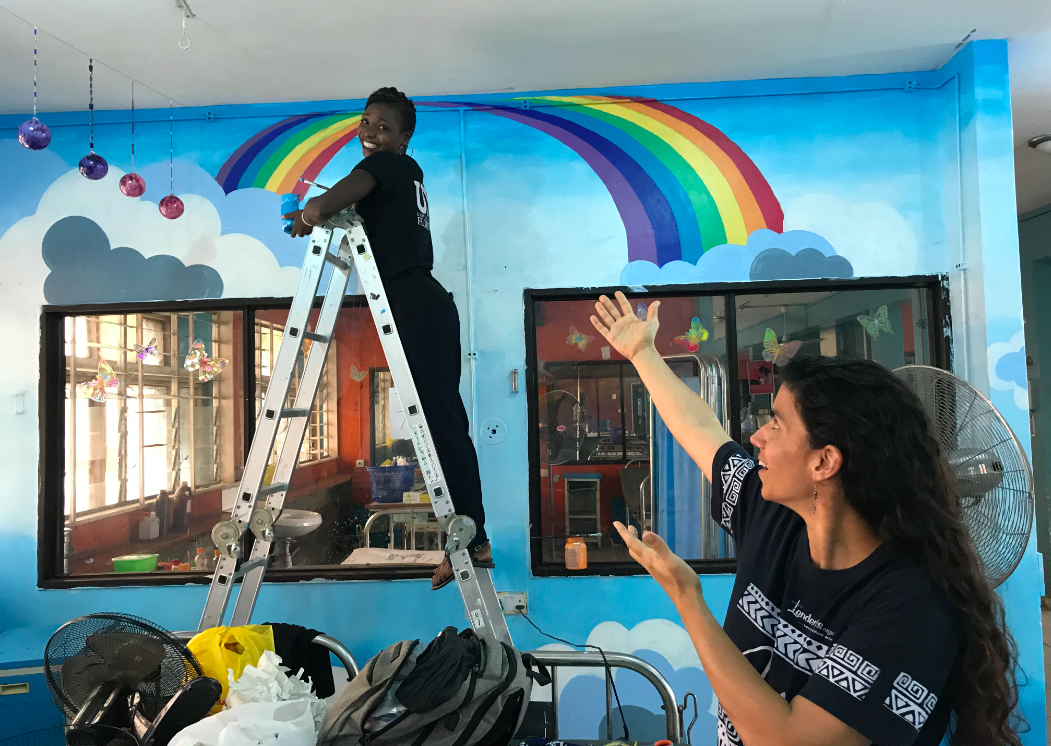 A woman points toward another woman who is painting a rainbow 