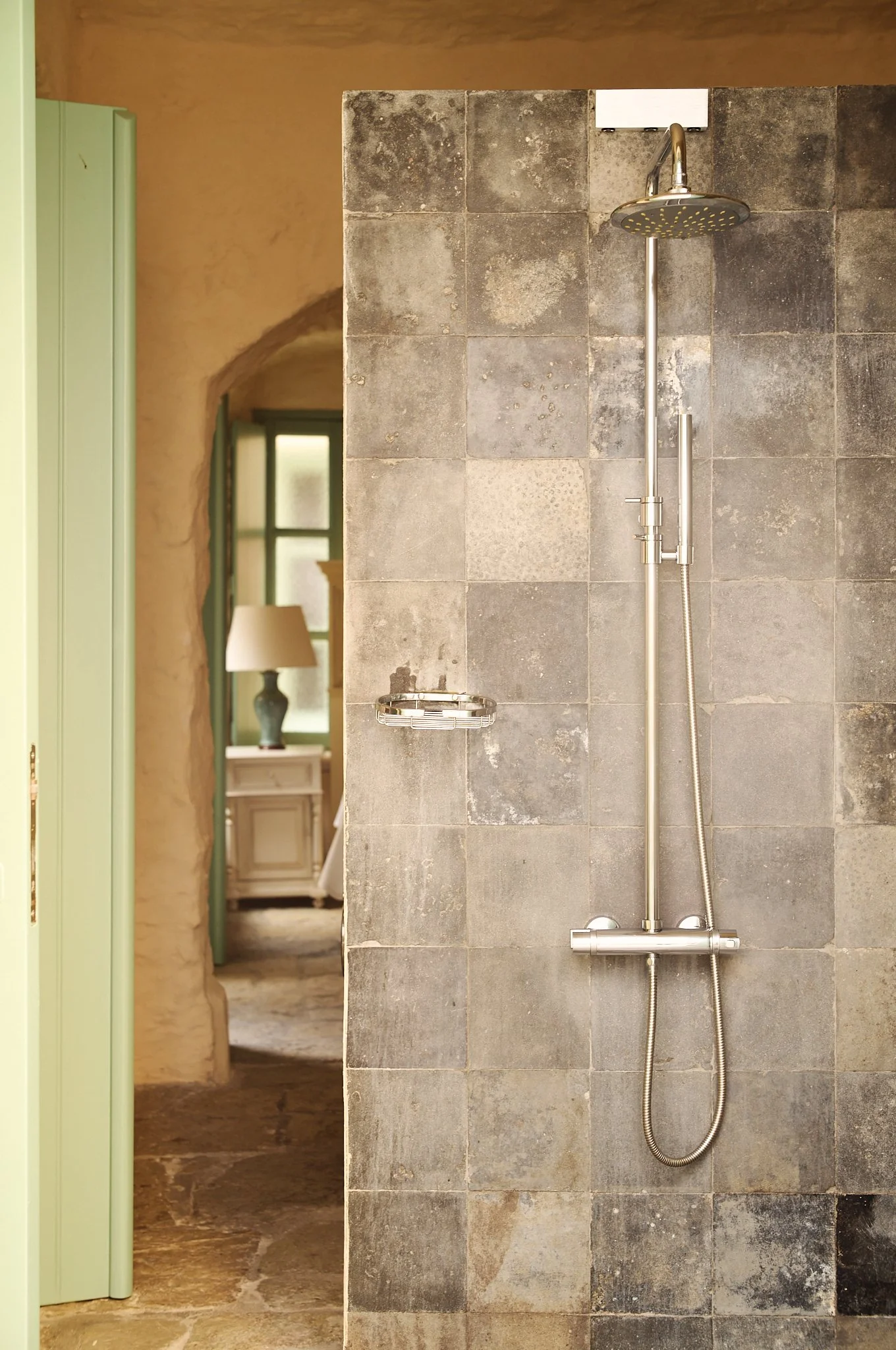 A shower area with a rain shower head, a handheld shower, and a small wire basket mounted on a gray tiled wall in a rustic space with a stone floor and a view into a room with a window and a lamp.