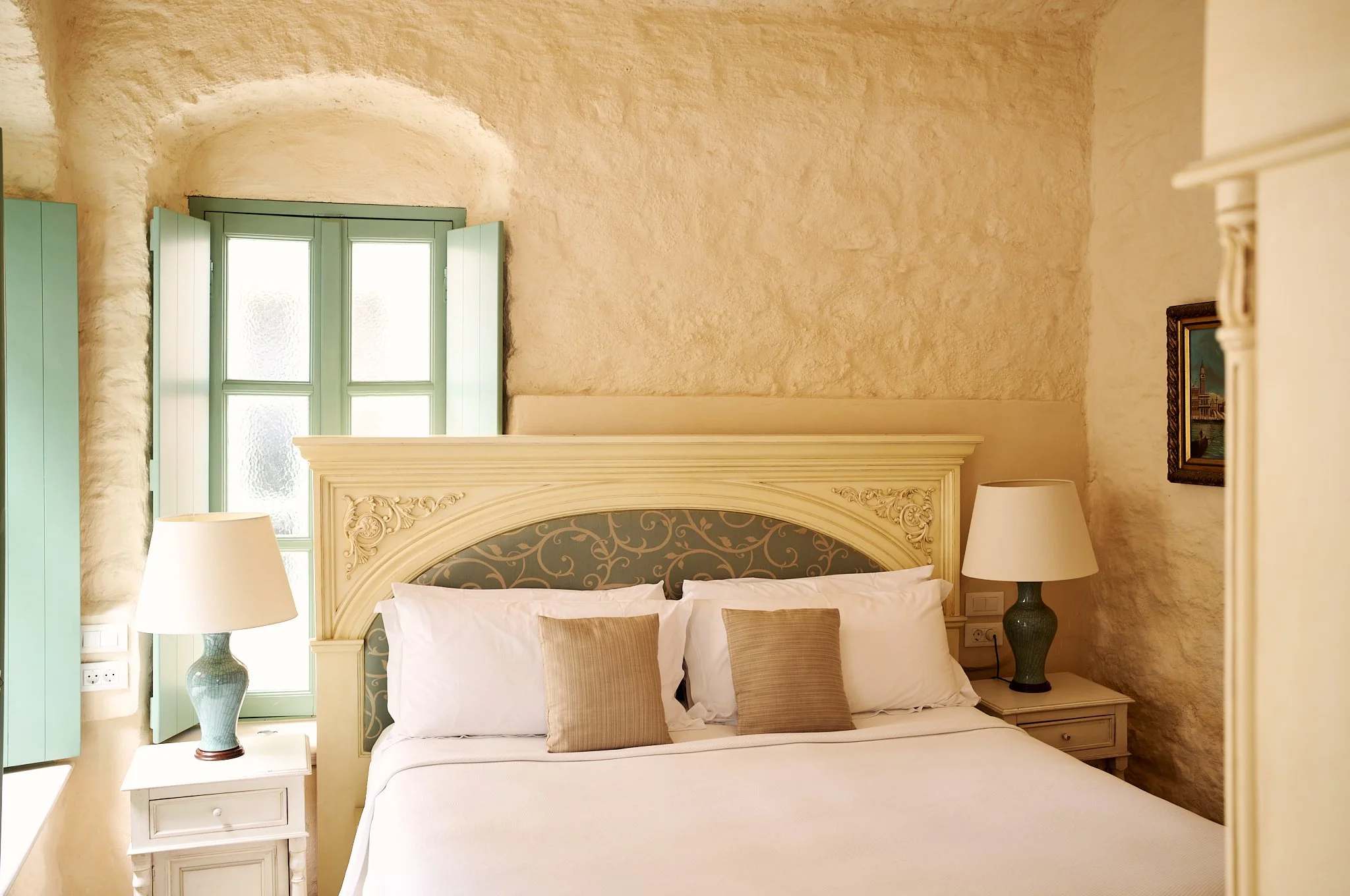 A bedroom with a large bed, beige pillows, a decorative headboard, two white lamps on dark green nightstands, a window with green shutters, and textured beige walls.