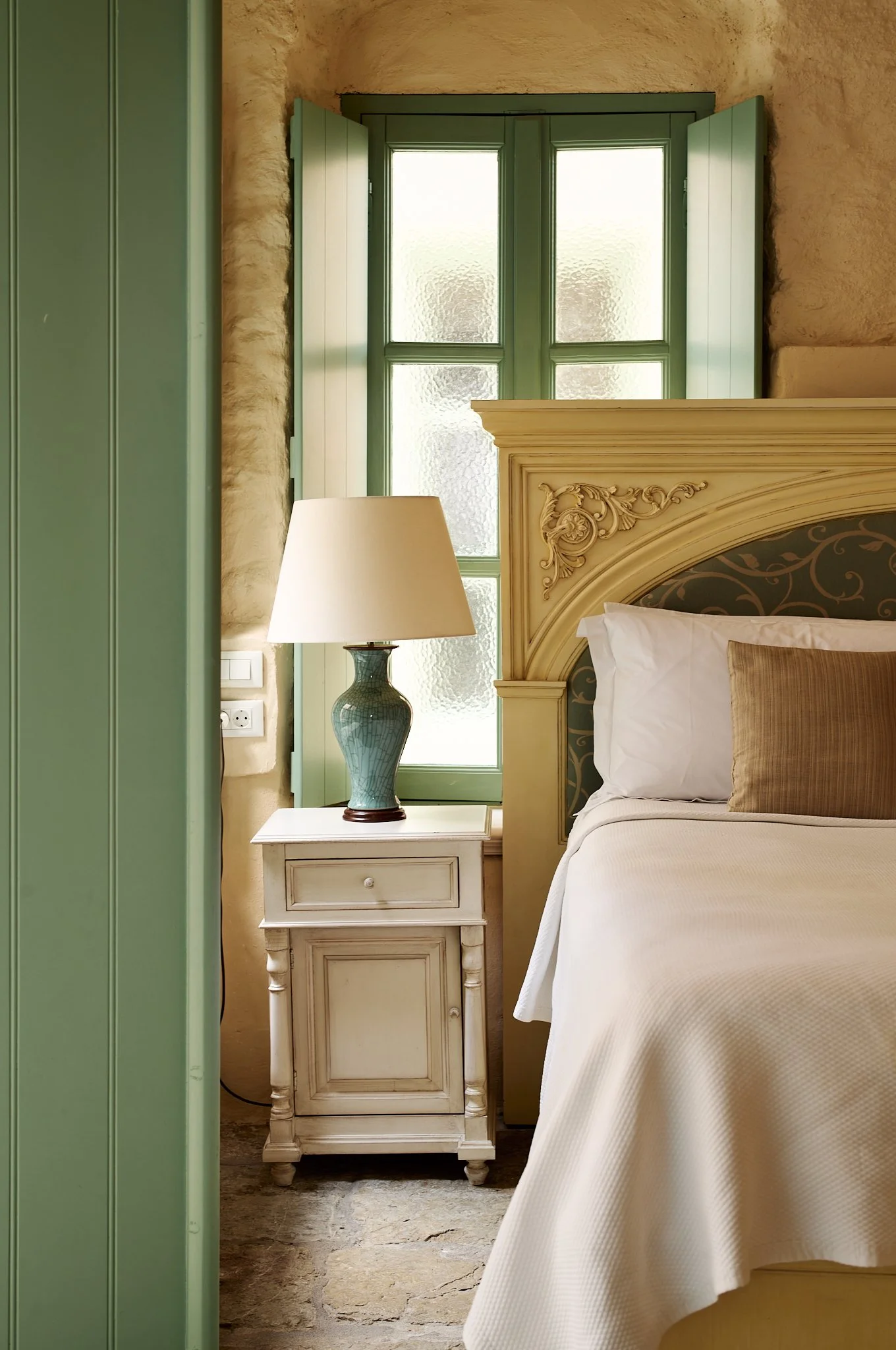 A bedroom corner with a ornate classic cream-colored headboard, white bedding, a beige pillow, a white bedside table with a light blue ceramic lamp, a green window with shutters, and a stone floor.