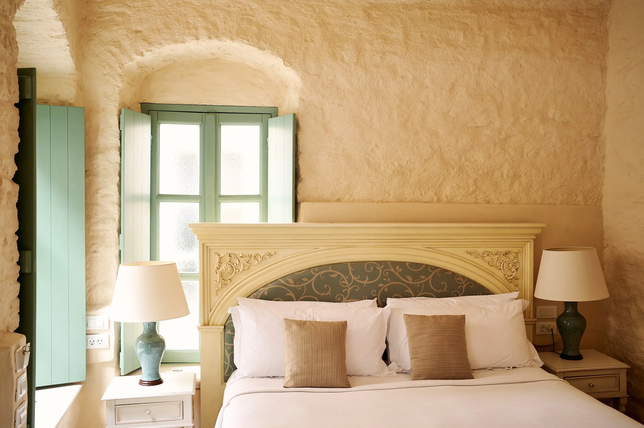 A cozy bedroom with an ornate cream-colored headboard, a pair of beige pillows, white bedding, and two matching lamps with cream-colored shades on nightstands. A window with green shutters lets in natural light, and the textured beige walls add warmt