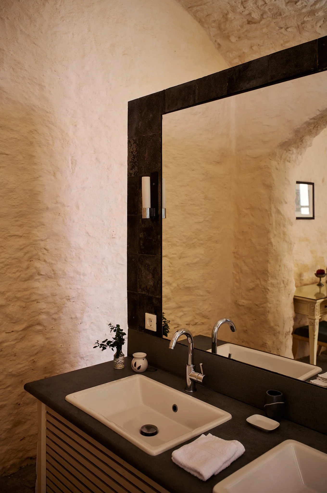 Bathroom vanity with double sinks, black countertop, and large mirror, decorated with small potted plant and white candle holder, against textured beige walls.