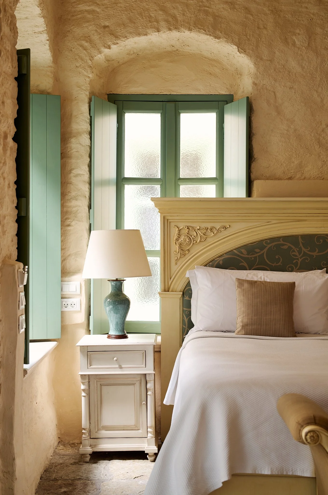 A cozy bedroom with a large window and green shutters, vintage cream-colored furniture, a white nightstand with a blue ceramic lamp, and a bed with white linens and beige pillows.
