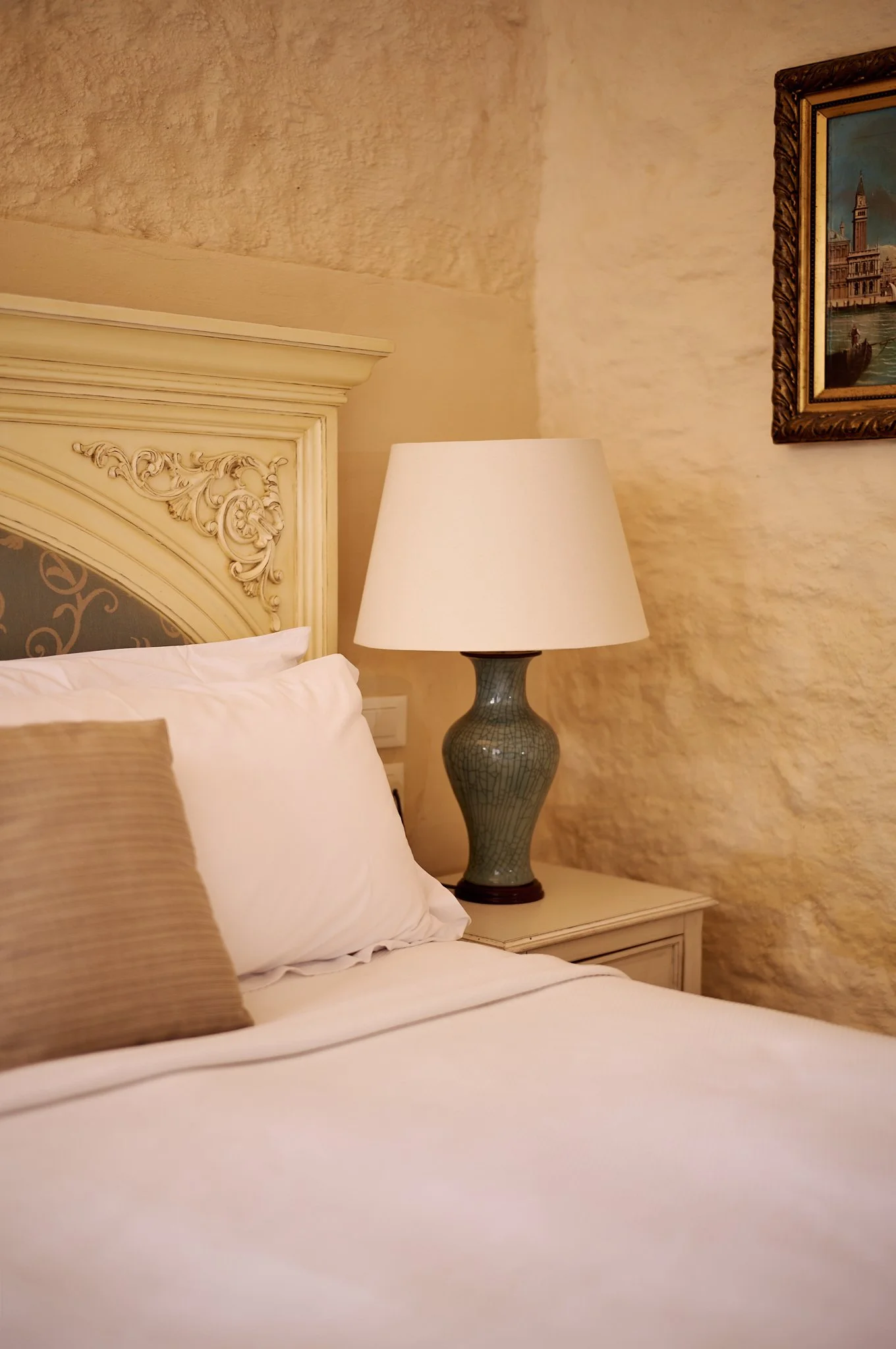 A bedroom with a cream-colored ornate headboard, a white bedside table, a table lamp, a white bed with pillows, a framed painting of Venice on the wall, and textured beige walls.